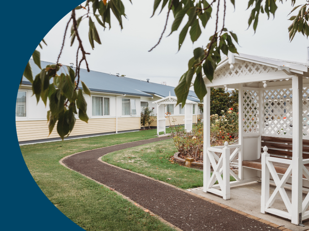 A garden path leading to a white gazebo with wooden benches, surrounded by greenery and a yellow house in the background.