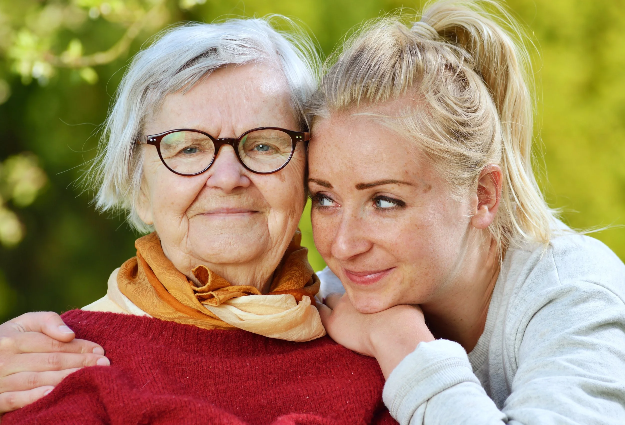 A young woman and an elderly woman embracing outdoors, with a blurred green background.