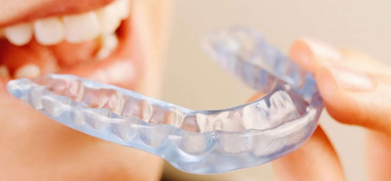 Close-up of a person holding a clear dental night guard in front of their open mouth, showing white teeth and gums.