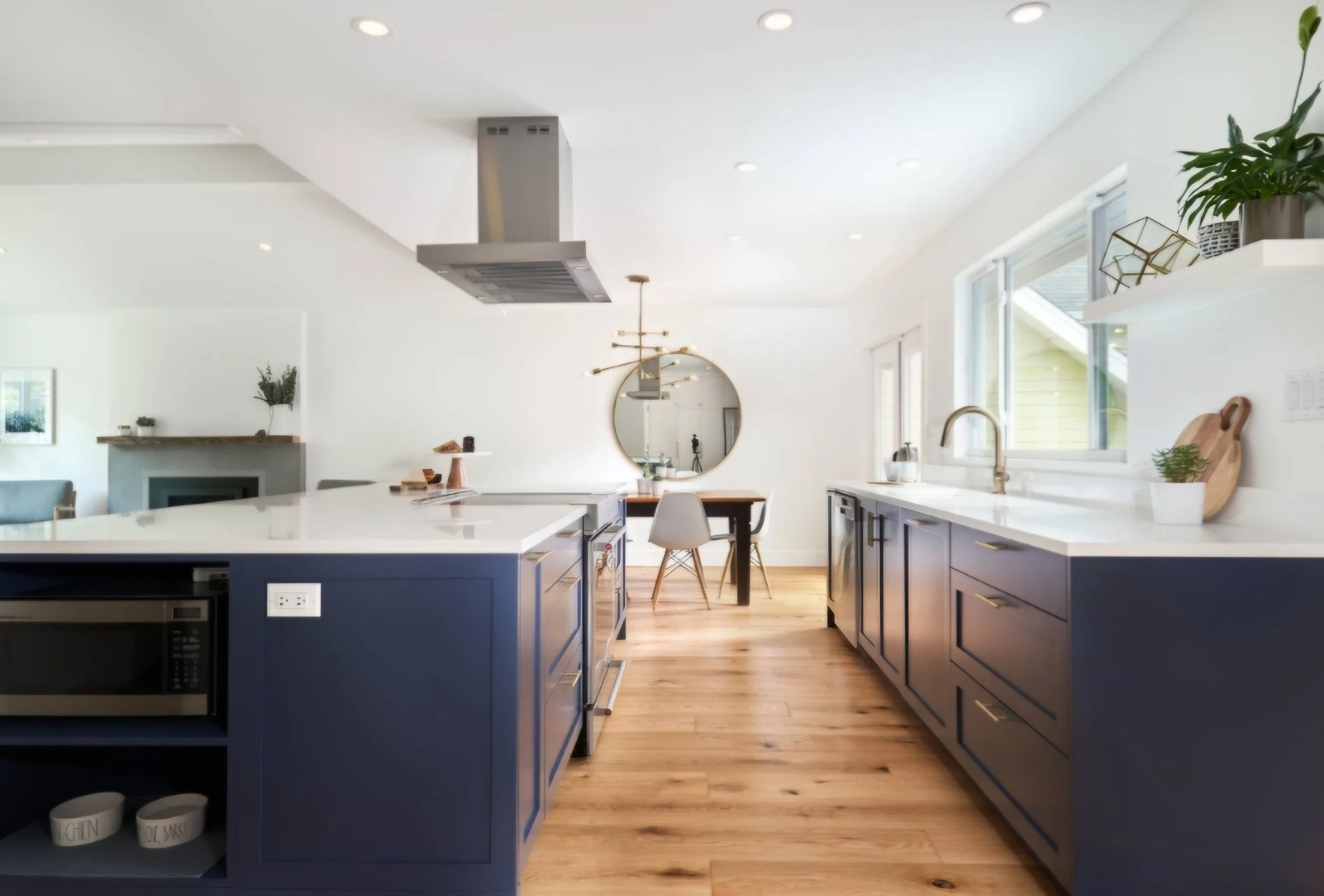 Modern kitchen with blue cabinets, white countertops, and hardwood flooring. A dining table with chairs is visible in the background, along with a round mirror on the wall, potted plants, and a large window.