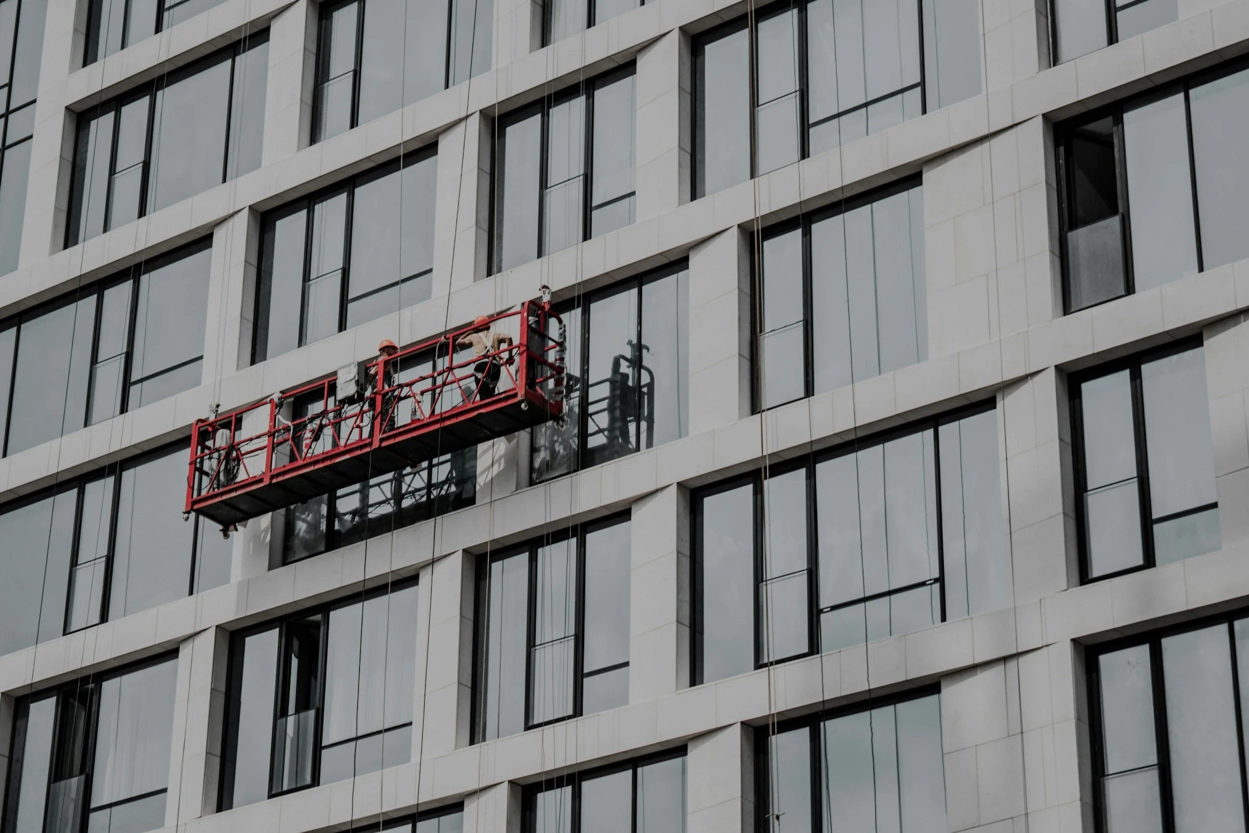 Workers on a suspended scaffold cleaning or maintaining the exterior of a tall modern building with large glass windows.