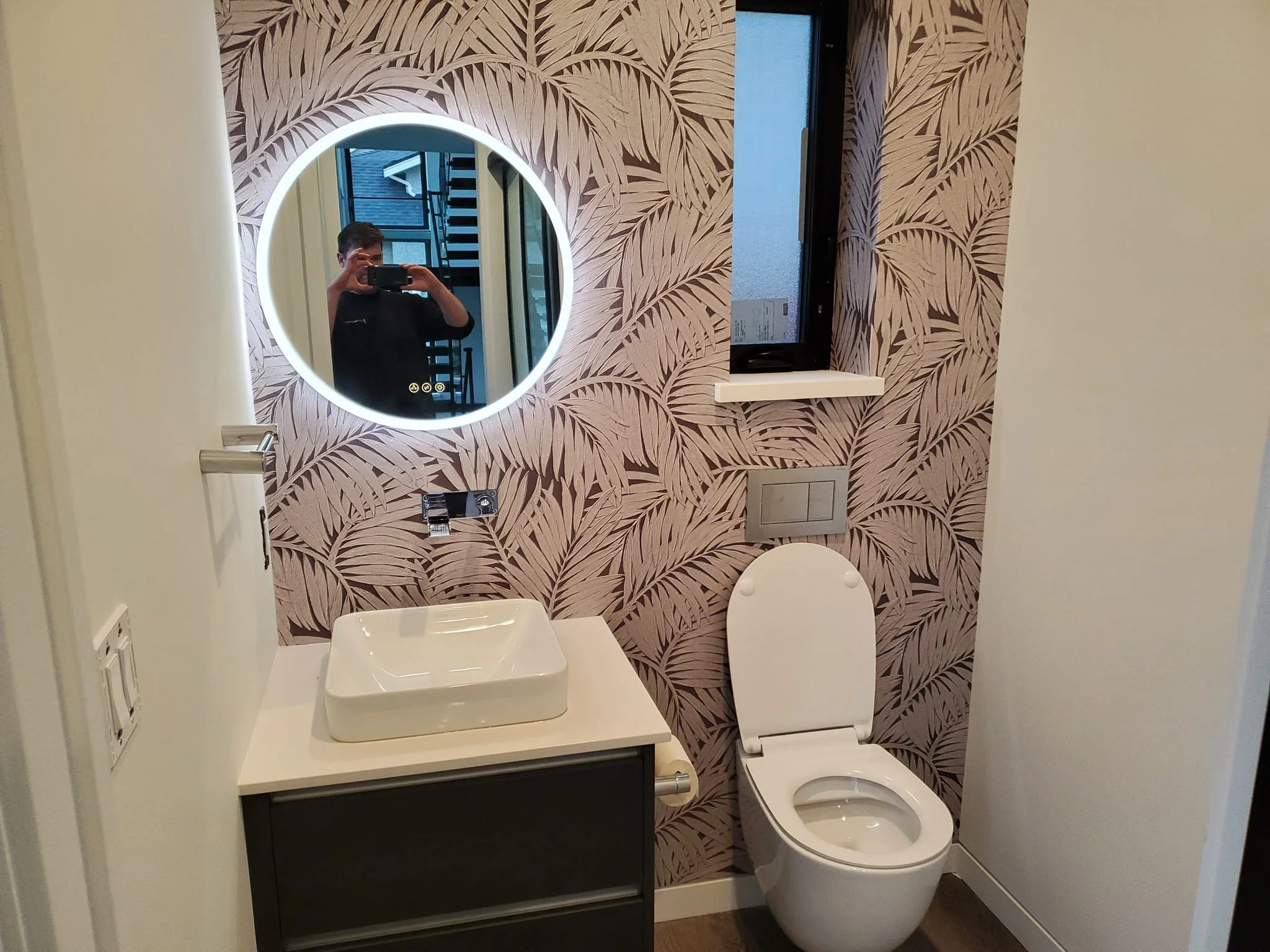 Modern bathroom with palm leaf patterned wallpaper, round illuminated mirror above a white vessel sink, white toilet, wall window, and a small shelf.