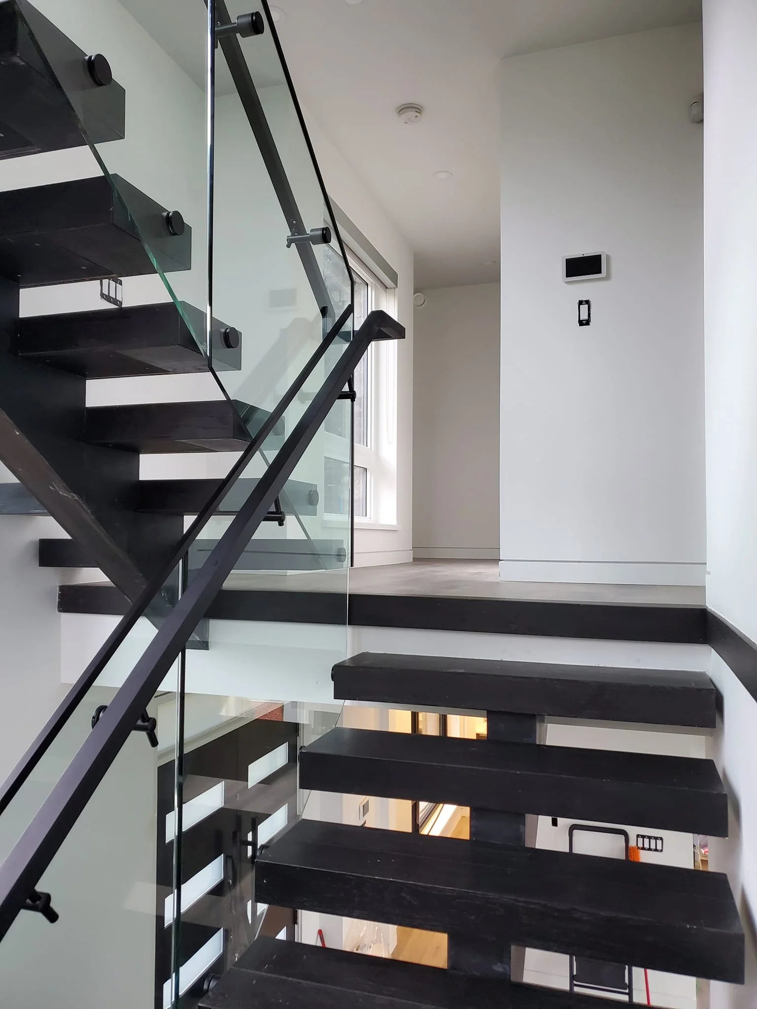 Interior view of a modern staircase with black steps and glass railing inside a house, with a window and white walls in the background.