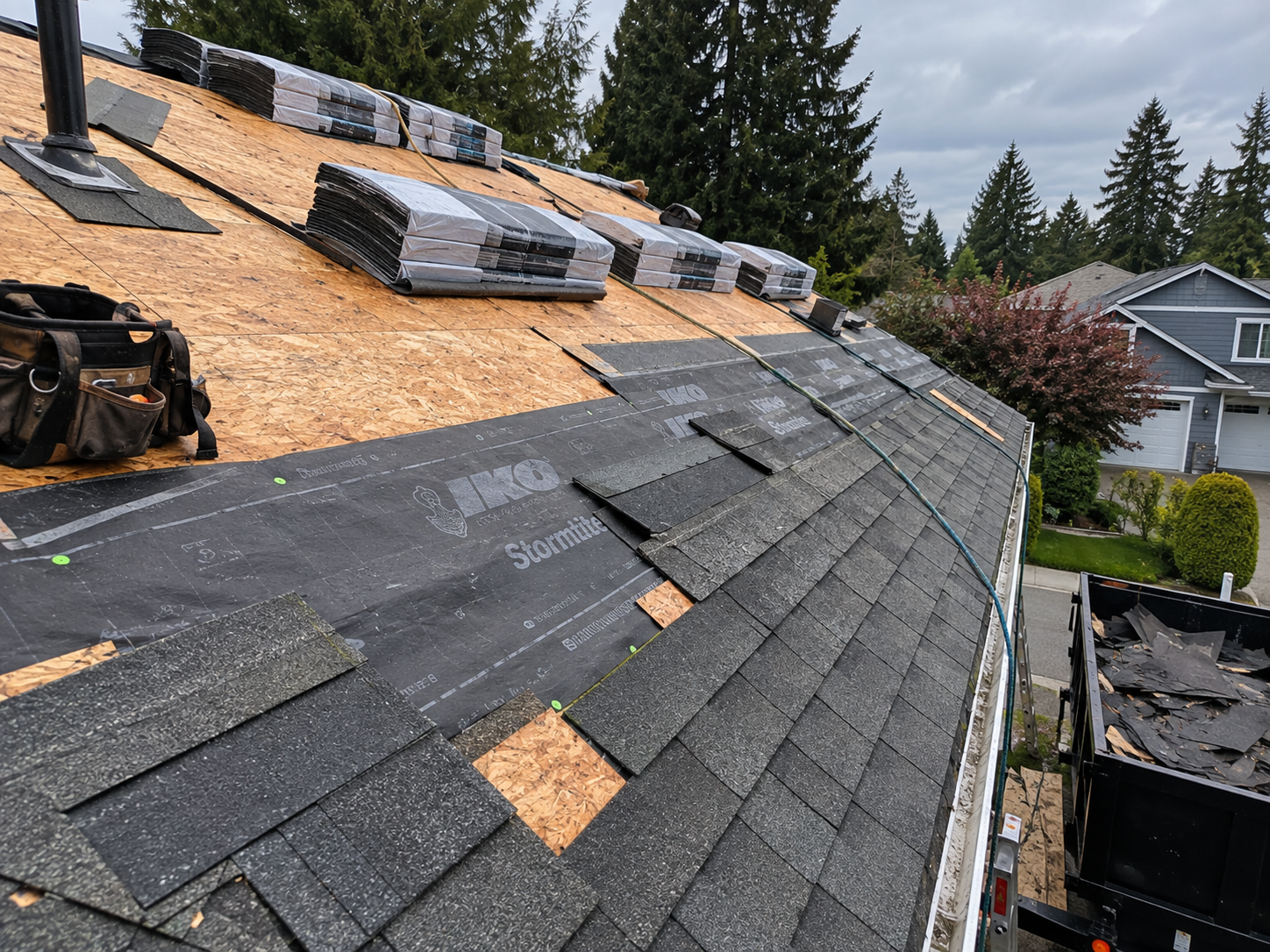 Roof under construction with asphalt shingles partially installed, bundles of roofing material stacked on the roof, and tools nearby, with a garage, trees, and a house in the background.