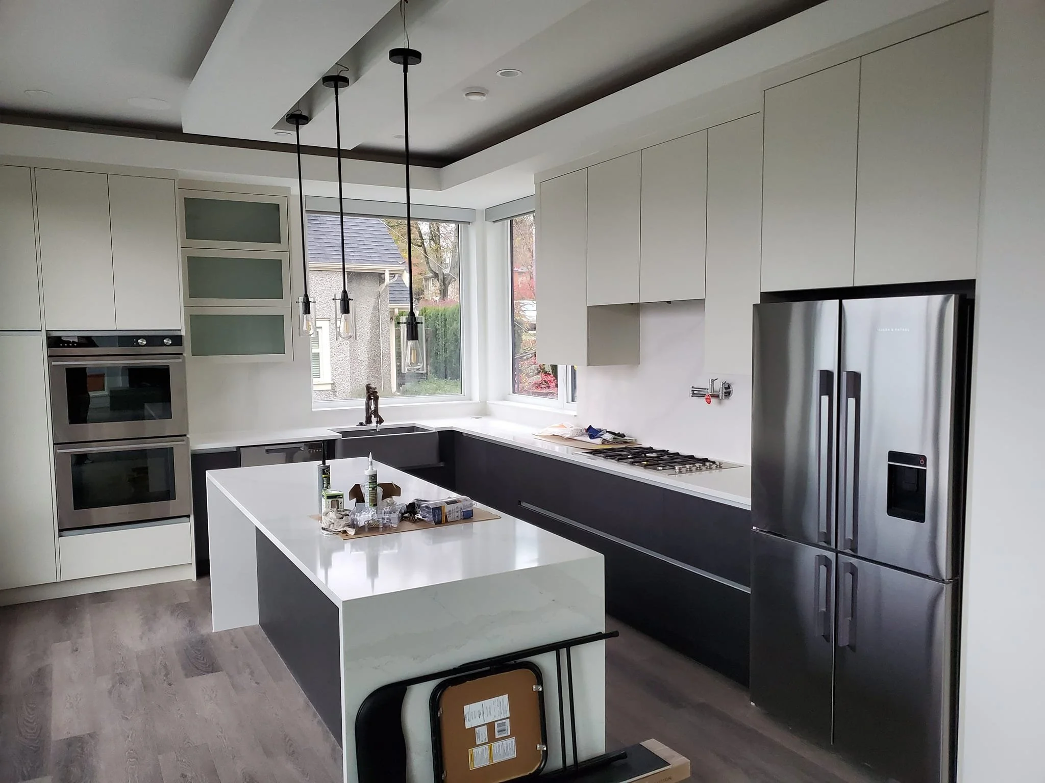 Modern kitchen with a white countertop island, stainless steel refrigerator, built-in double oven, and a window showing an outdoor view. Hanging pendant lights are above the island.