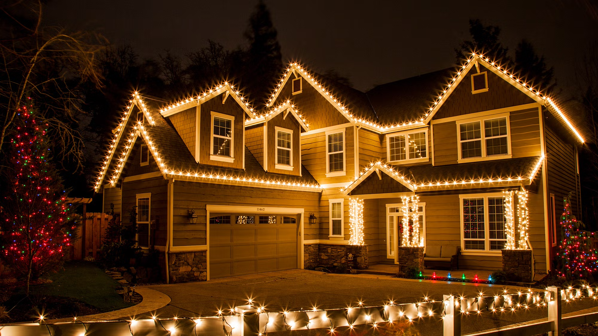 A house decorated with Christmas lights at night, featuring white string lights outlining the roof, windows, and porch columns, with Christmas trees decorated with multicolored lights on both sides.
