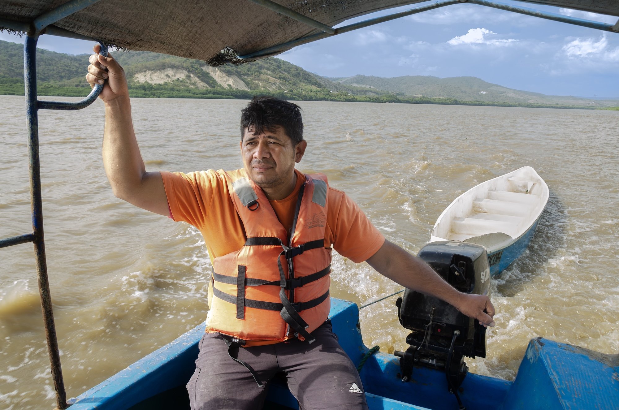 Hombre en una lancha con chaleco salvavidas, sosteniendo el timón, en un río con paisaje montañoso al fondo.