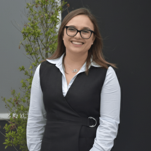 Mujer sonriendo con gafas, blanca y con cabello castaño, vestida con blusa blanca y chaleco negro, de pie frente a un fondo gris con plantas