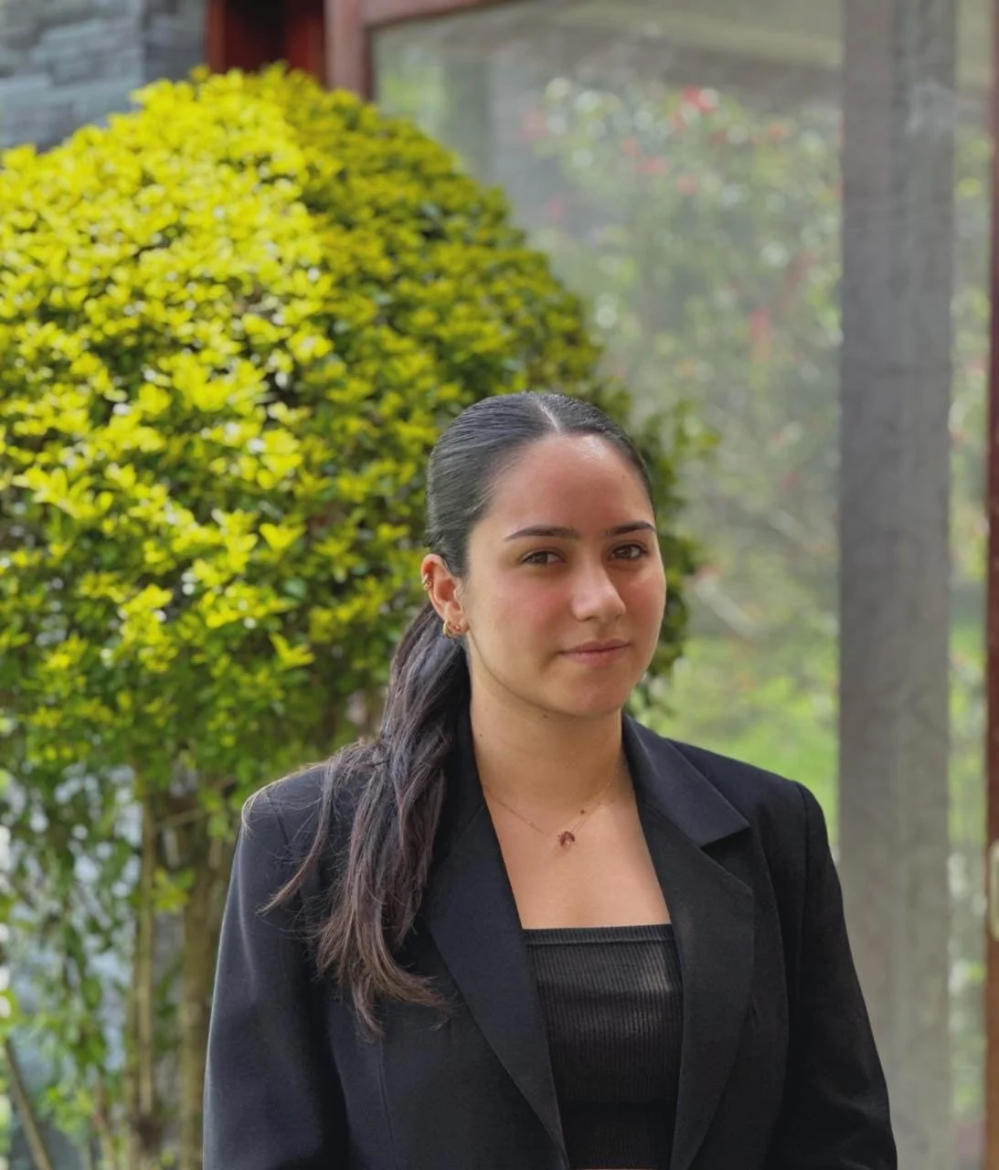 Joven mujer con cabello oscuro en una coleta, lleva un blazer negro y una blusa negra, posando al aire libre con arbustos verdes y un fondo difuso con árboles y una estructura de vidrio.