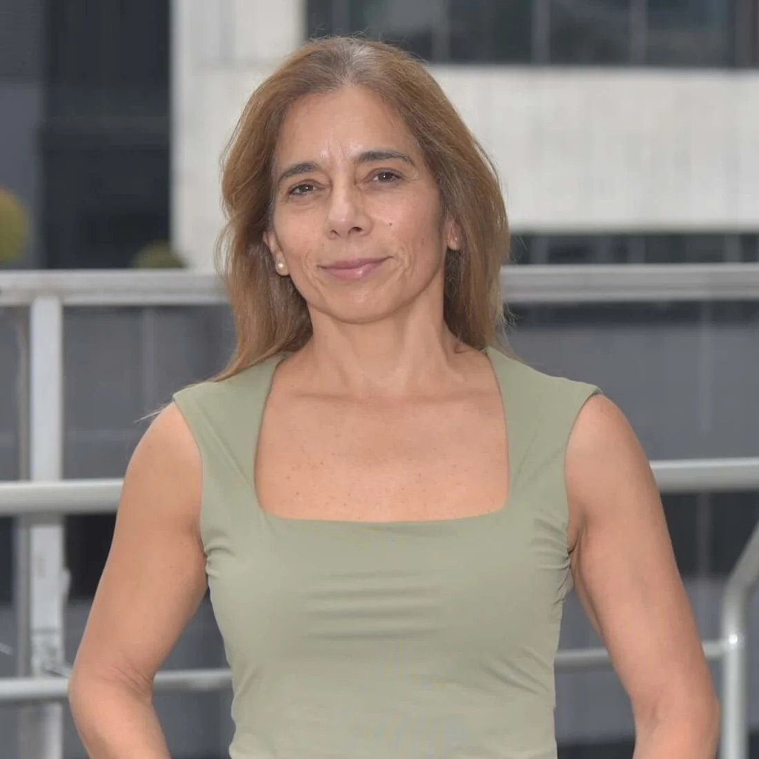 Mujer joven con cabello liso y rubio claro, usa una blusa de tirantes gris y está al aire libre con edificios de fondo.