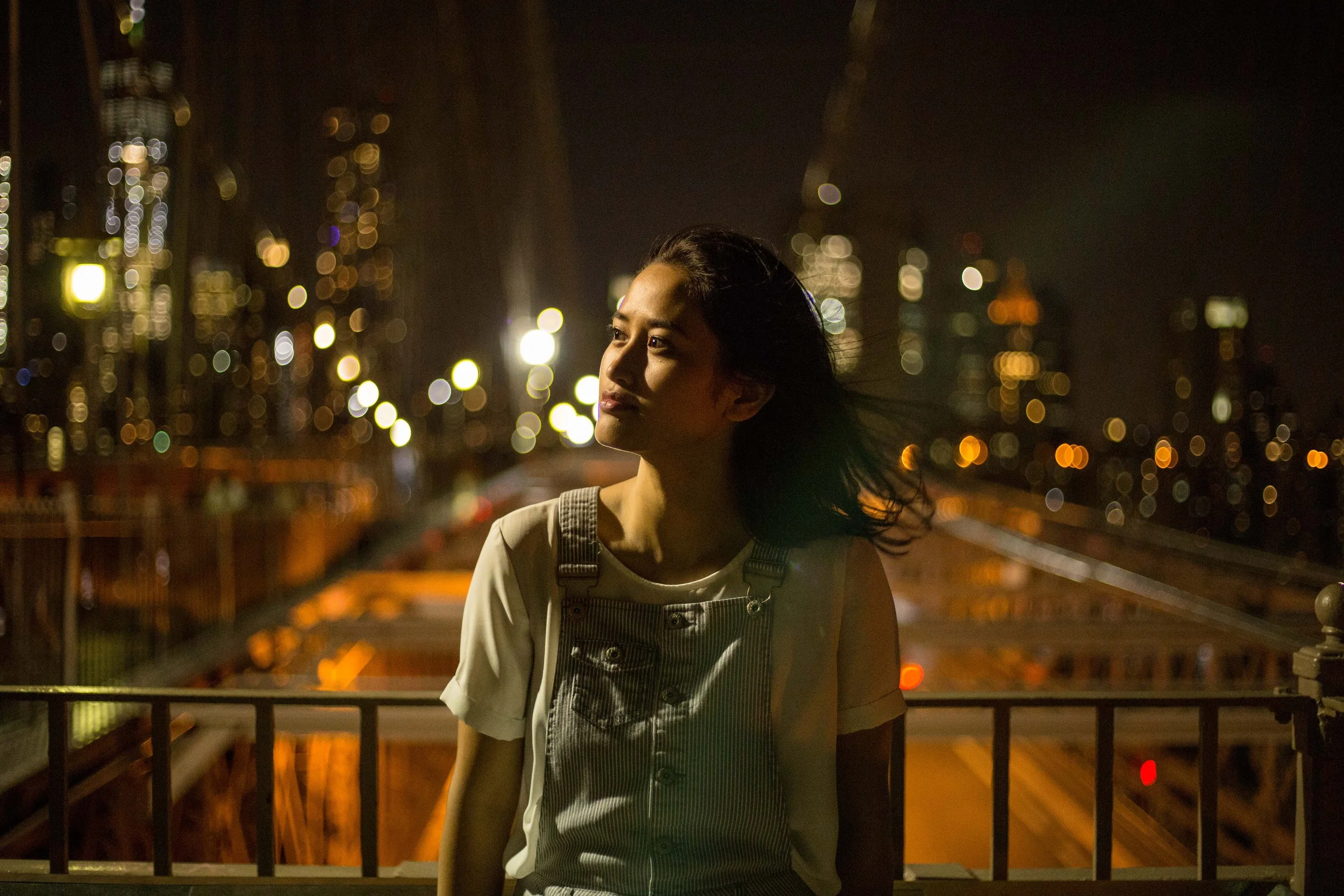 A woman with long black hair standing on a bridge at night, with city lights and skyscrapers in the blurred background.