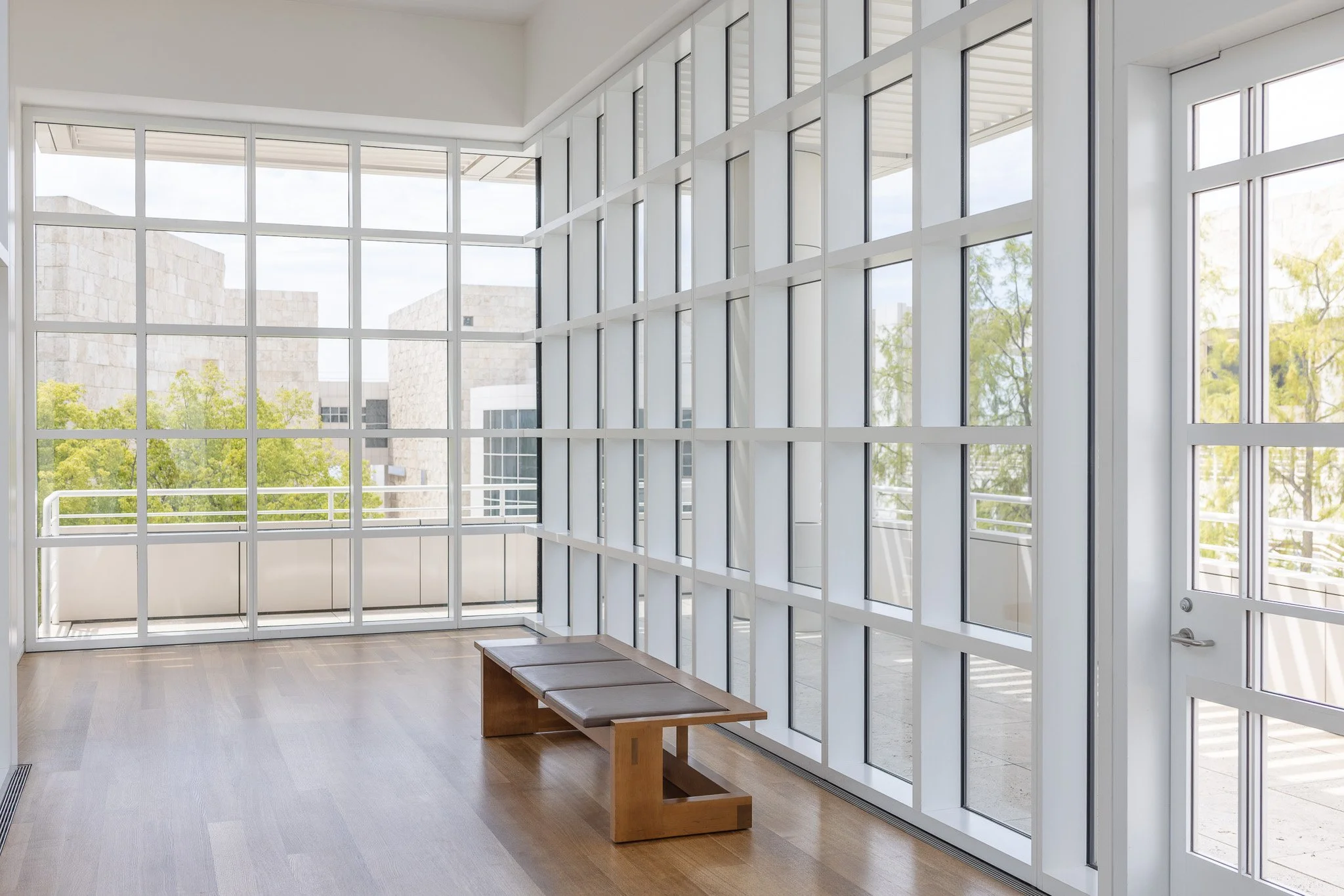 Interior of a modern room with large glass windows, a wooden bench, and a view of trees and neighboring buildings outside.