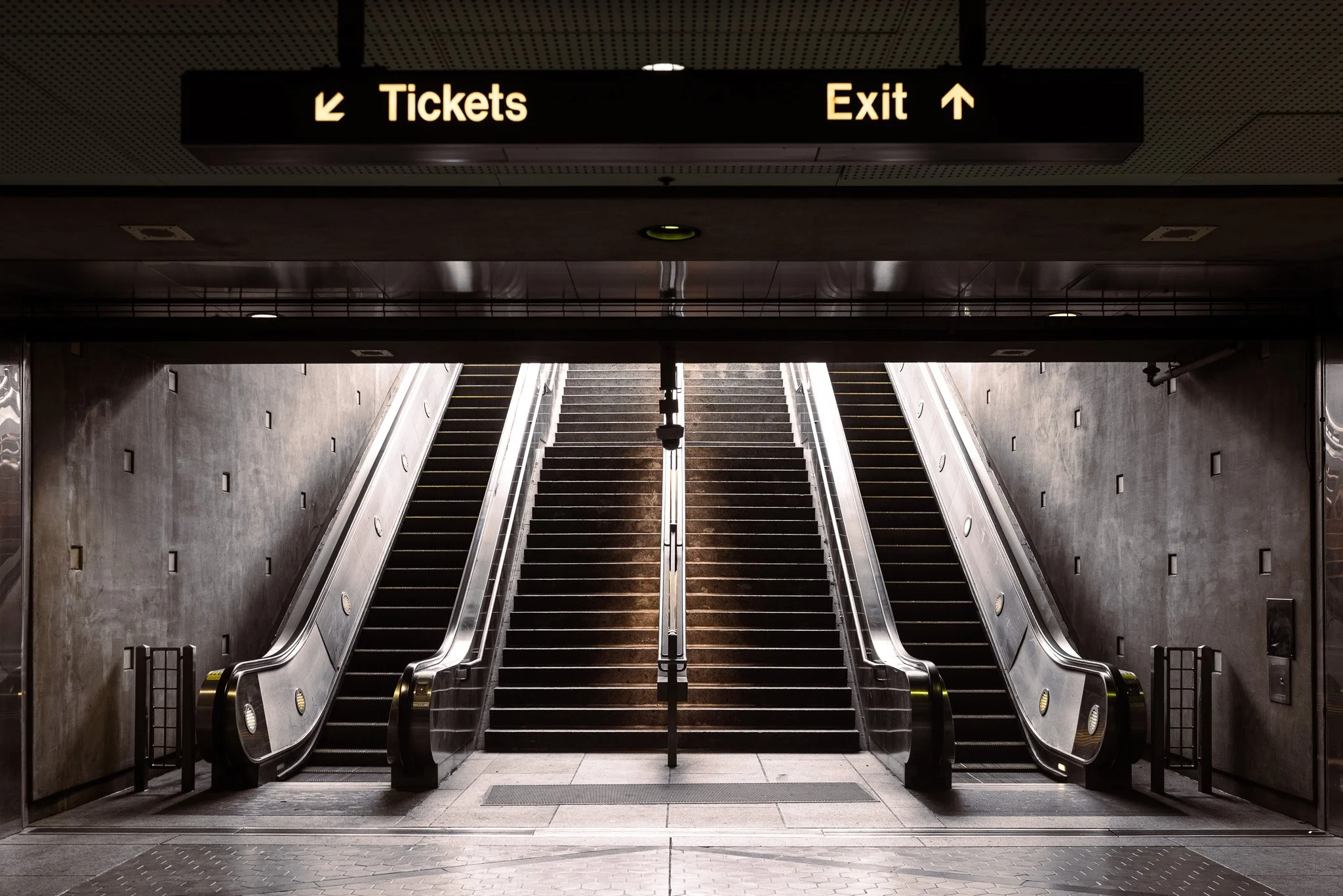 Inside view of an underground transit station with escalators leading to the upper level, with a sign overhead indicating 'Tickets' to the left and 'Exit' straight ahead.