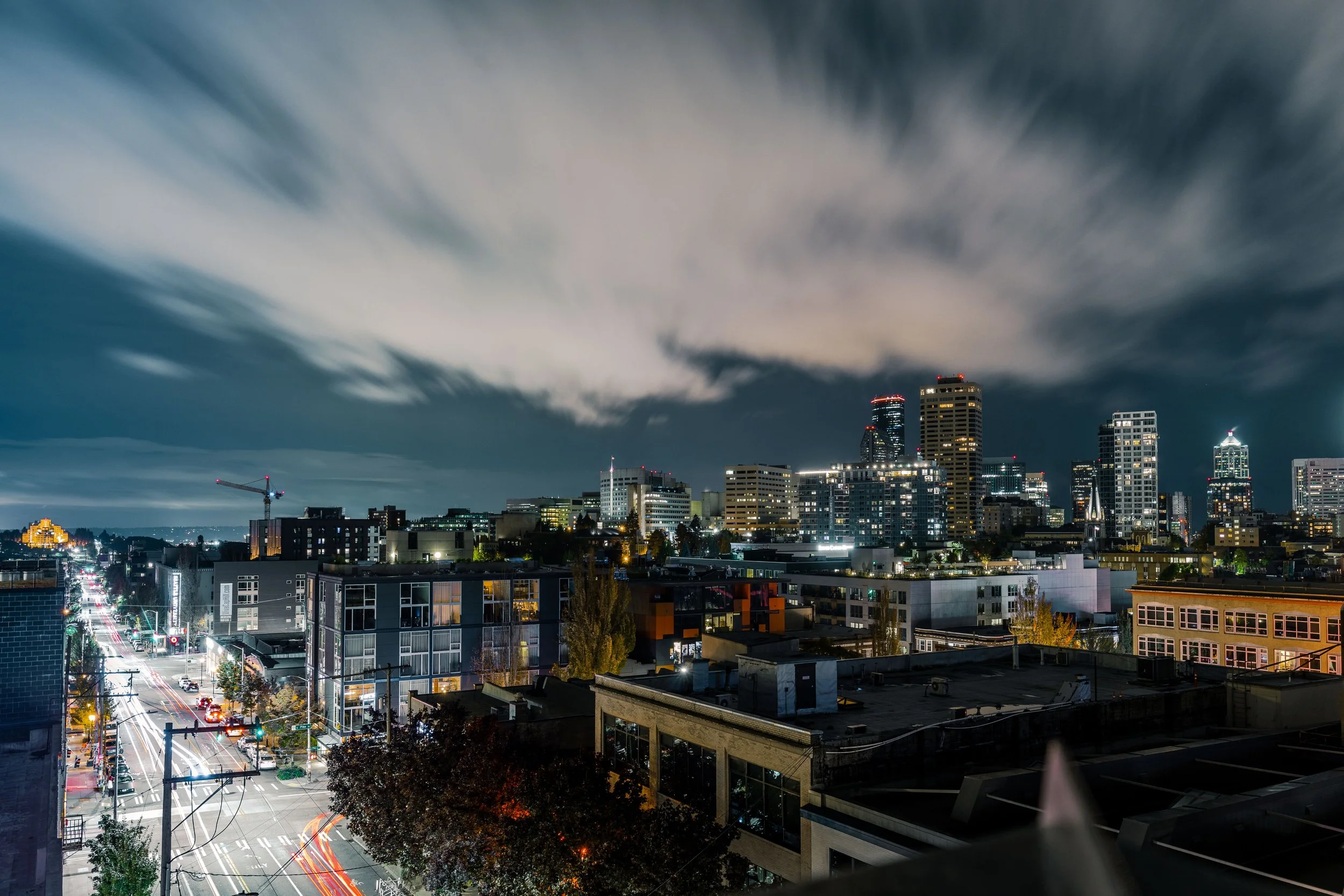 Night view of a city skyline with tall buildings and illuminated streets below, some clouds are visible in the sky.