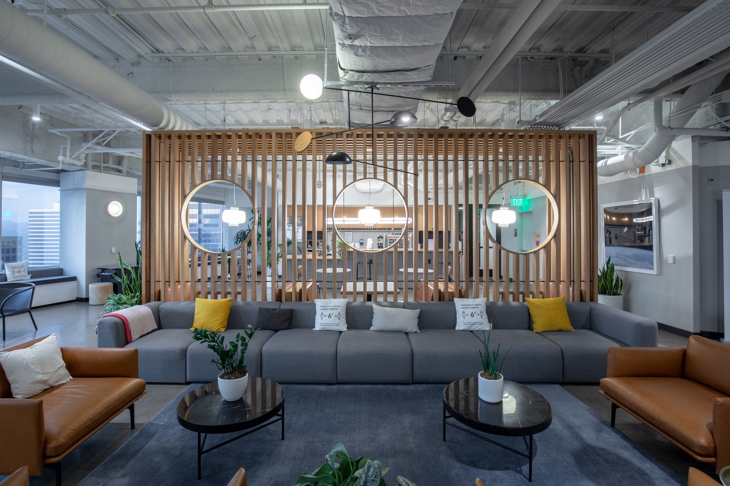 Modern office lounge with gray sofa, brown leather chairs, coffee tables with plants, decorative wooden slat partition with circular mirrors, and large windows in the background.
