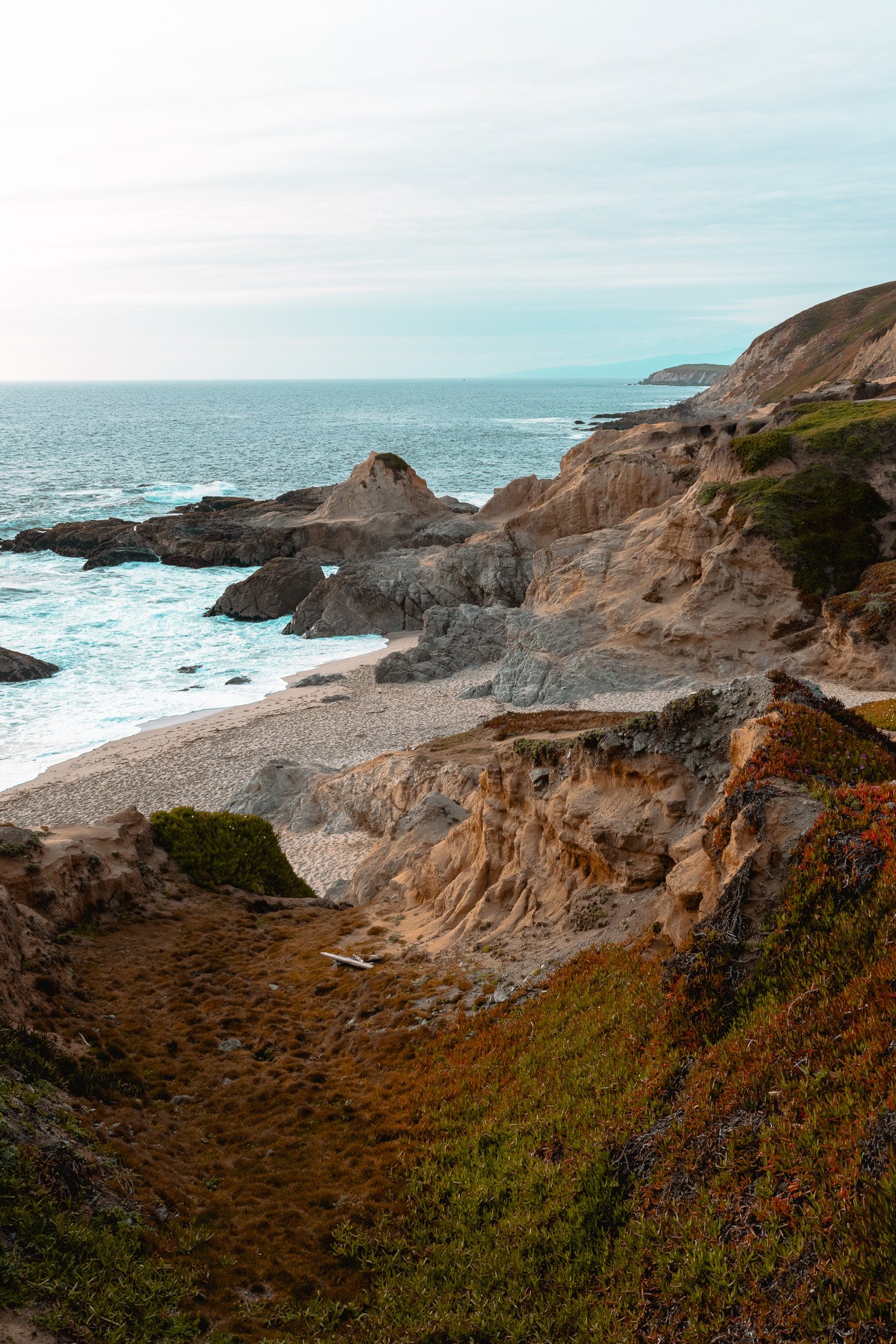 Coastal landscape with rocky cliffs, sandy beach, and ocean waves under a cloudy sky.
