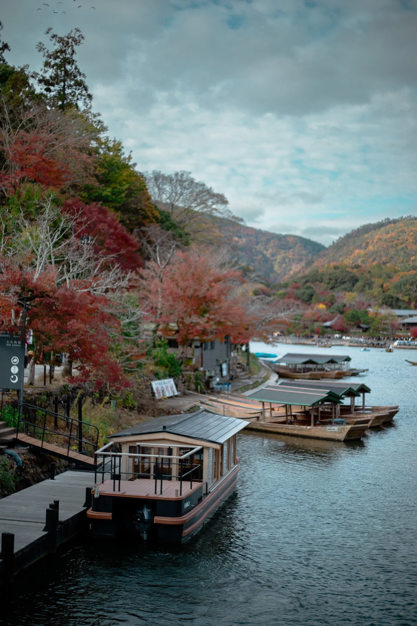 A lakeside scene with boats docked at a small pier, surrounded by colorful autumn trees and hills in the background, under a cloudy sky.