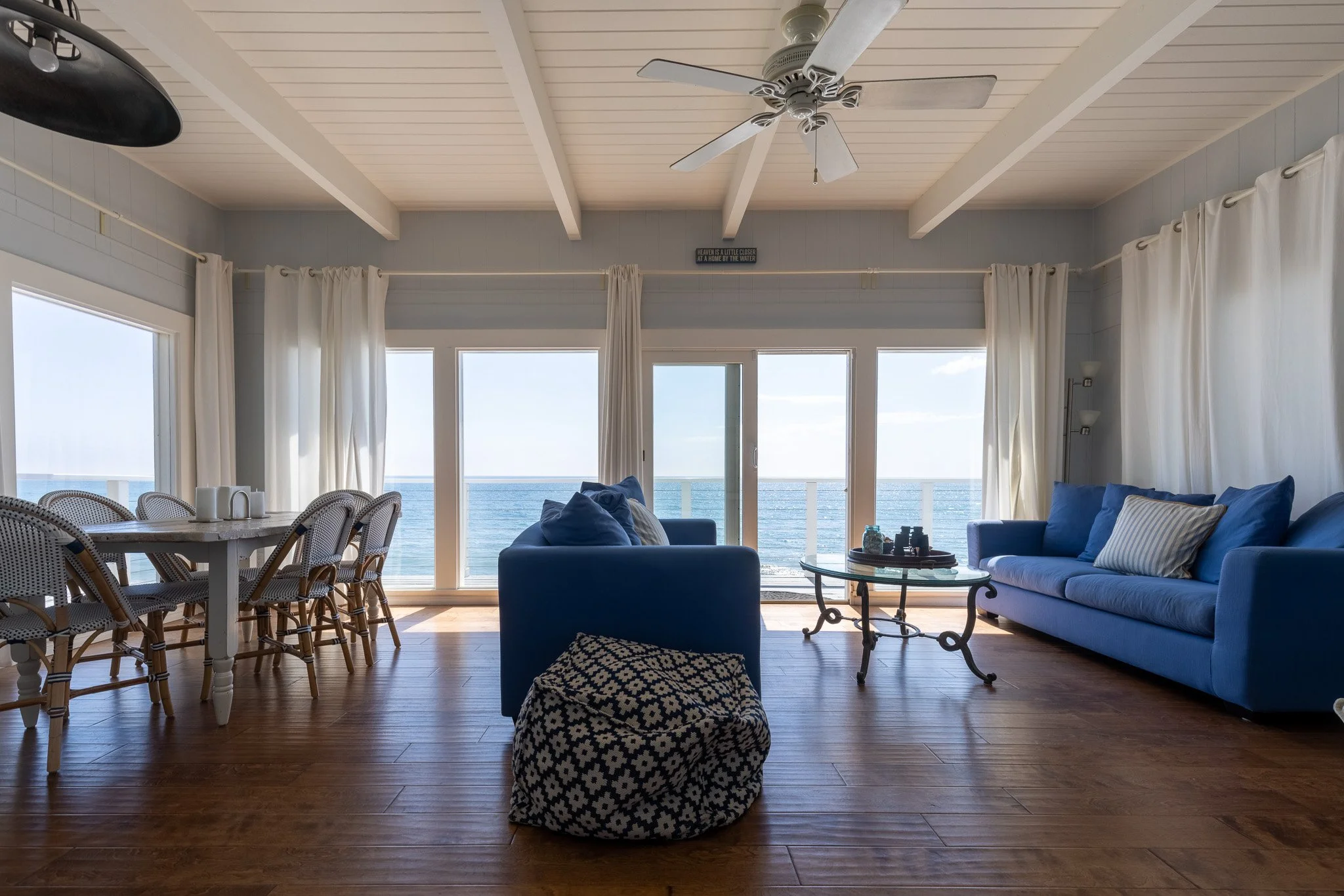 Living room with ocean view, blue sofas, dining table, white curtains, wooden floor, ceiling fan, and wall lamps.