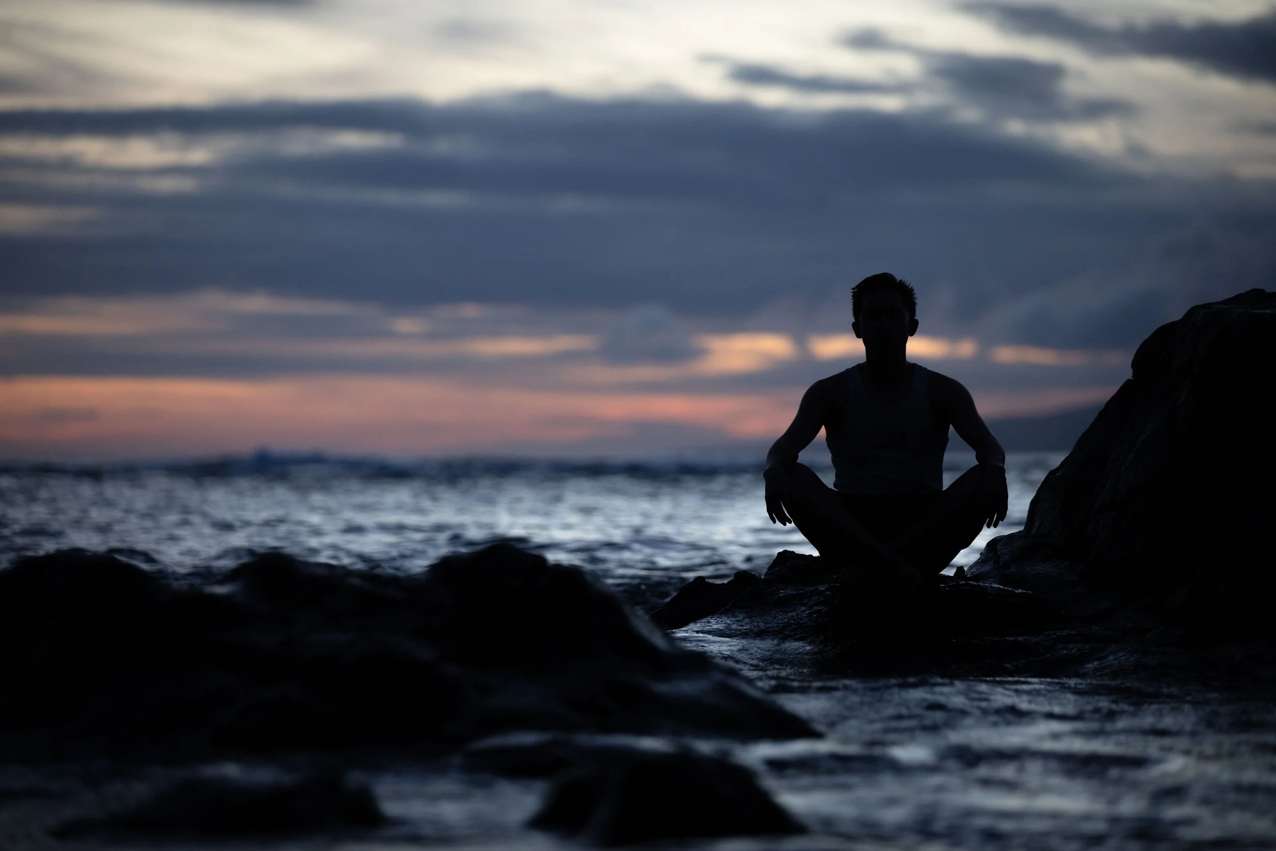 Silhouette of a person sitting cross-legged on rocks at the beach during sunset with a cloudy sky.