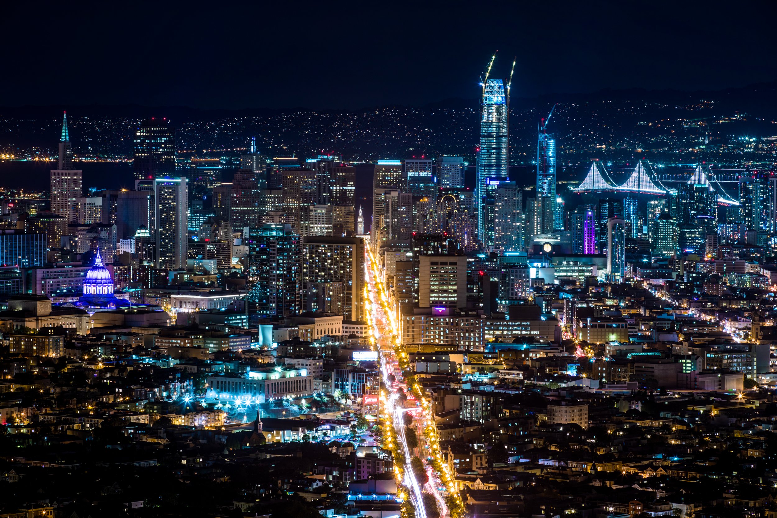 Nighttime aerial view of San Francisco skyline with illuminated buildings, including the Salesforce Tower and numerous skyscrapers, as well as bridges and city streets with bright lights.