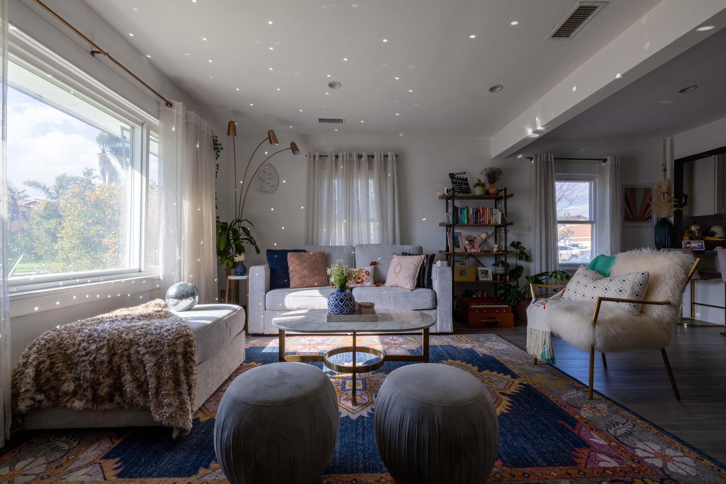 Bright living room with large window, white sofa with colorful pillows, modern armchair with fluffy throw, oval marble coffee table, two black ottomans, hardwood floors, and decorative area rug, decorated with plants, books, and art.
