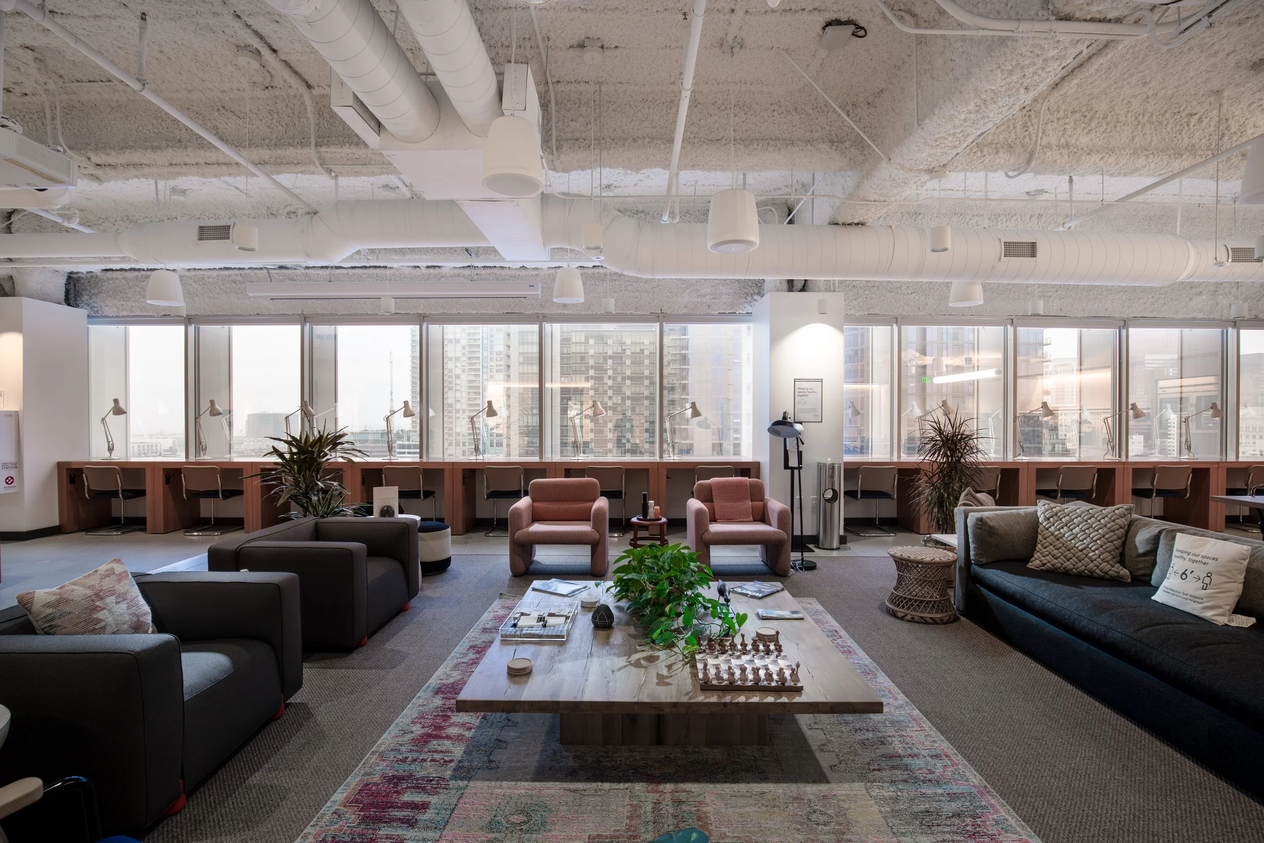 An office lounge area with a large wooden coffee table, surrounded by various armchairs and sofas, large windows with city views, fluorescent lighting, plants, and a carpet with a colorful pattern.