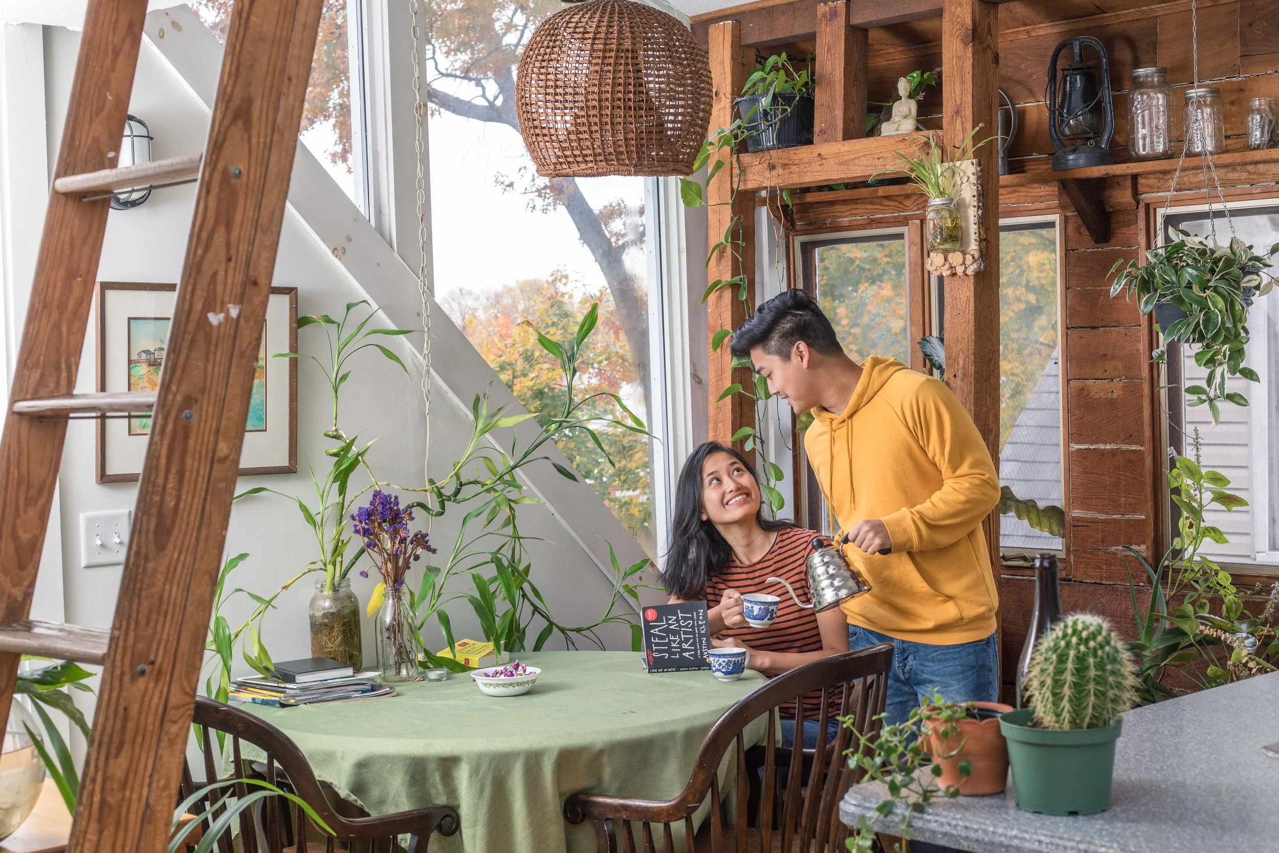 A young man and woman sharing a moment in a cozy, plant-filled dining space with wooden walls and large windows, with the man pouring tea into a cup held by the woman.