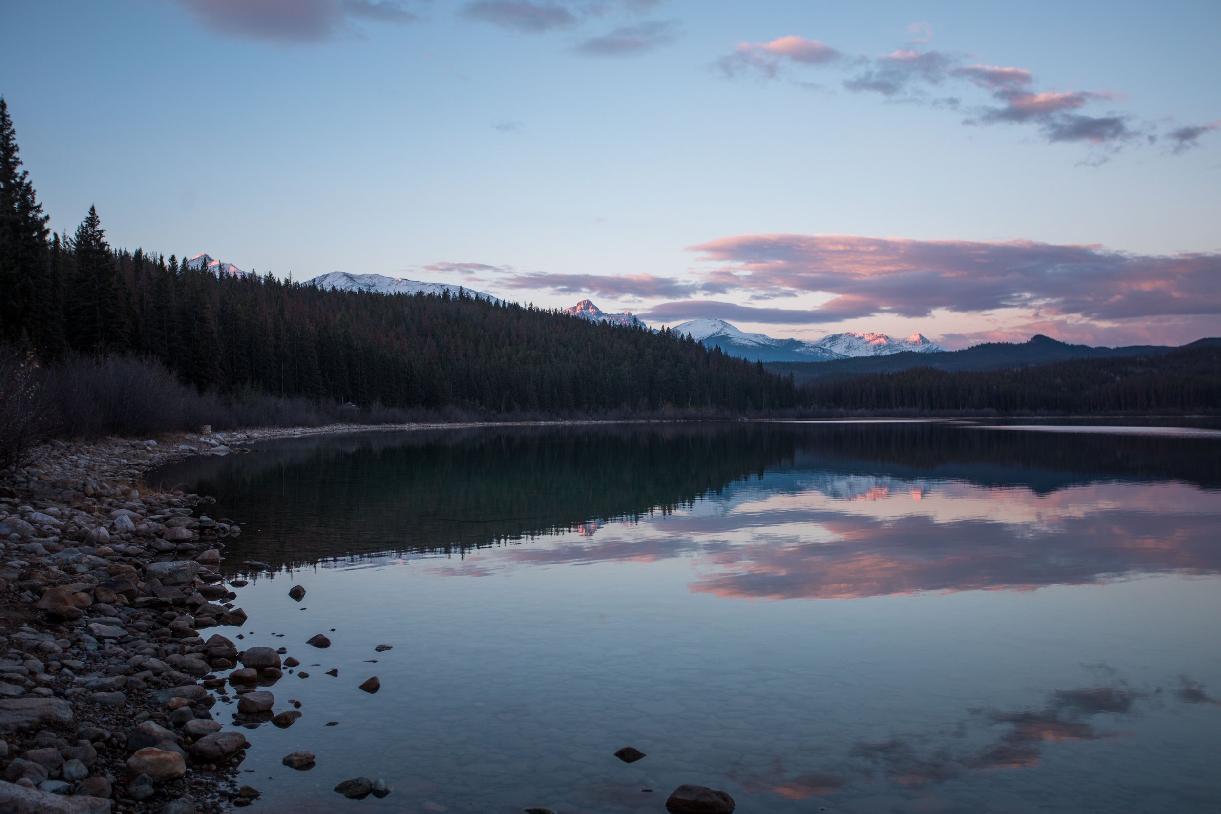Scenic view of a calm mountain lake at dusk with snow-capped peaks in the background, reflected in the water, and a rocky shoreline in the foreground.