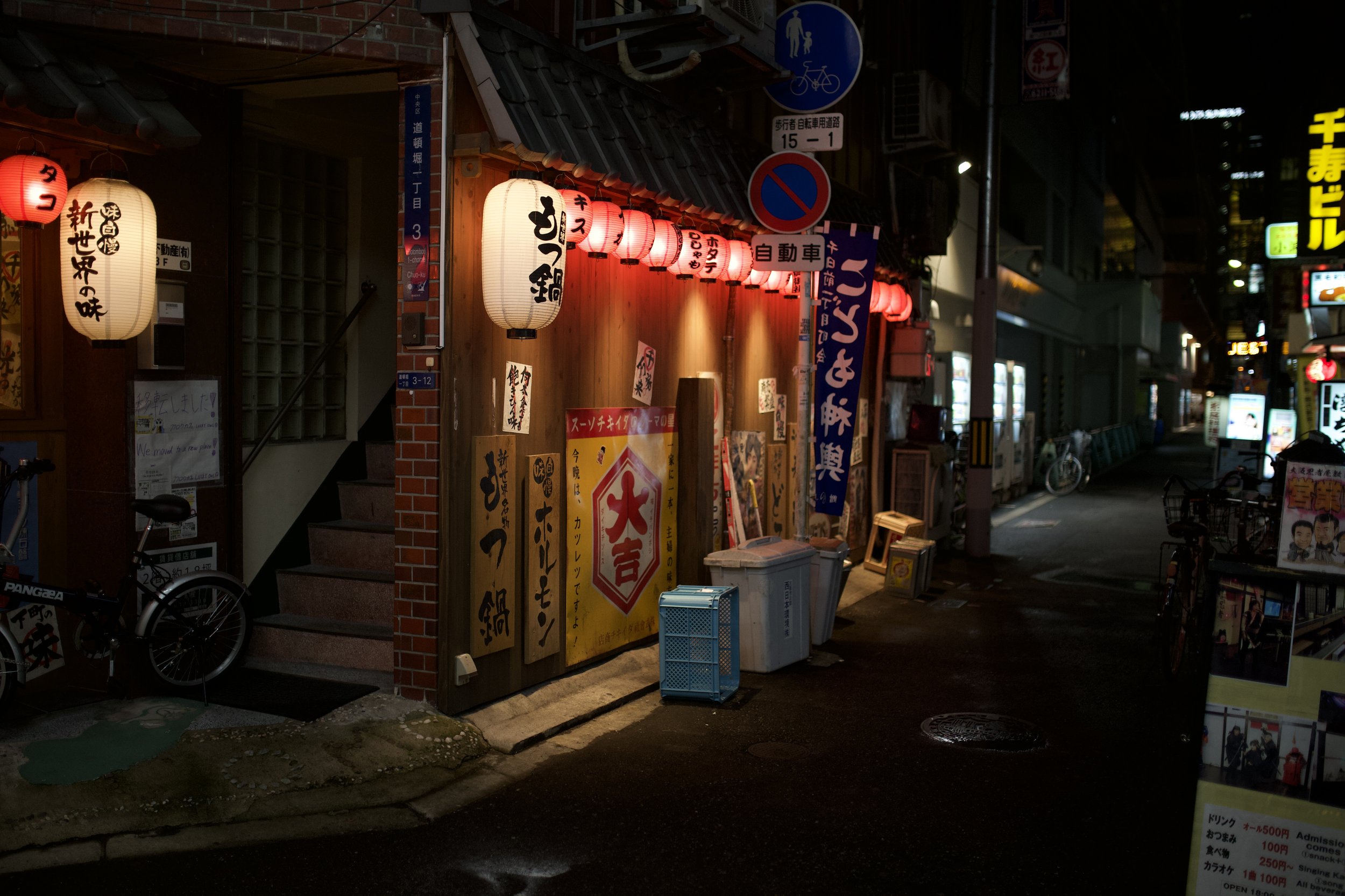 Street scene at night with a Japanese restaurant illuminated by lanterns, signs in Japanese, bicycles parked outside, and advertising boards on the sidewalk.