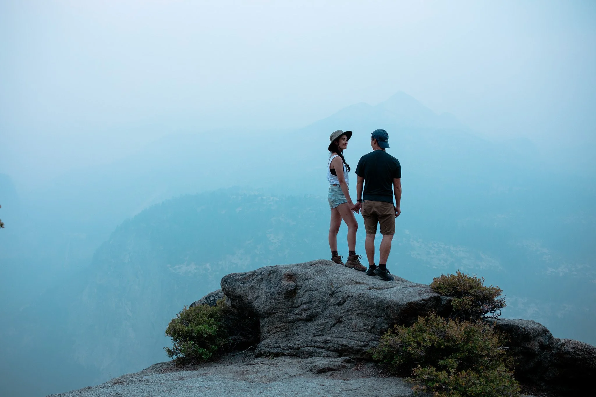 A couple holding hands on a large rock at a mountain overlook, with a hazy mountain range in the background.