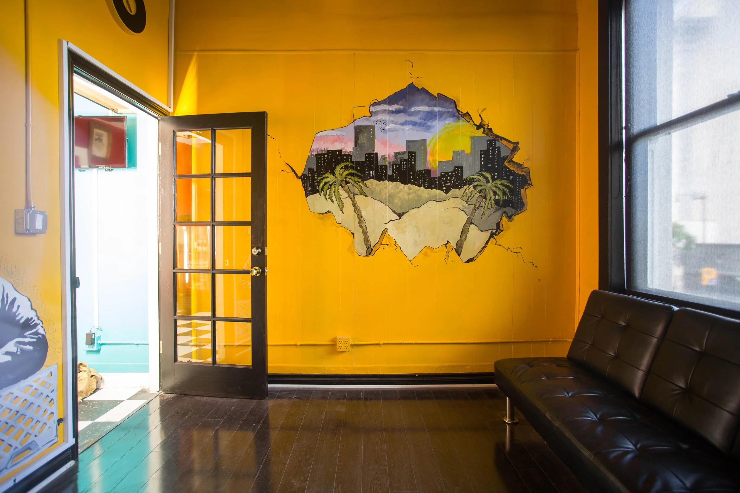 Interior of a room with a yellow wall featuring a mural of a city skyline and palm trees, a black door with glass panes, a window with a shade, and a black leather bench.