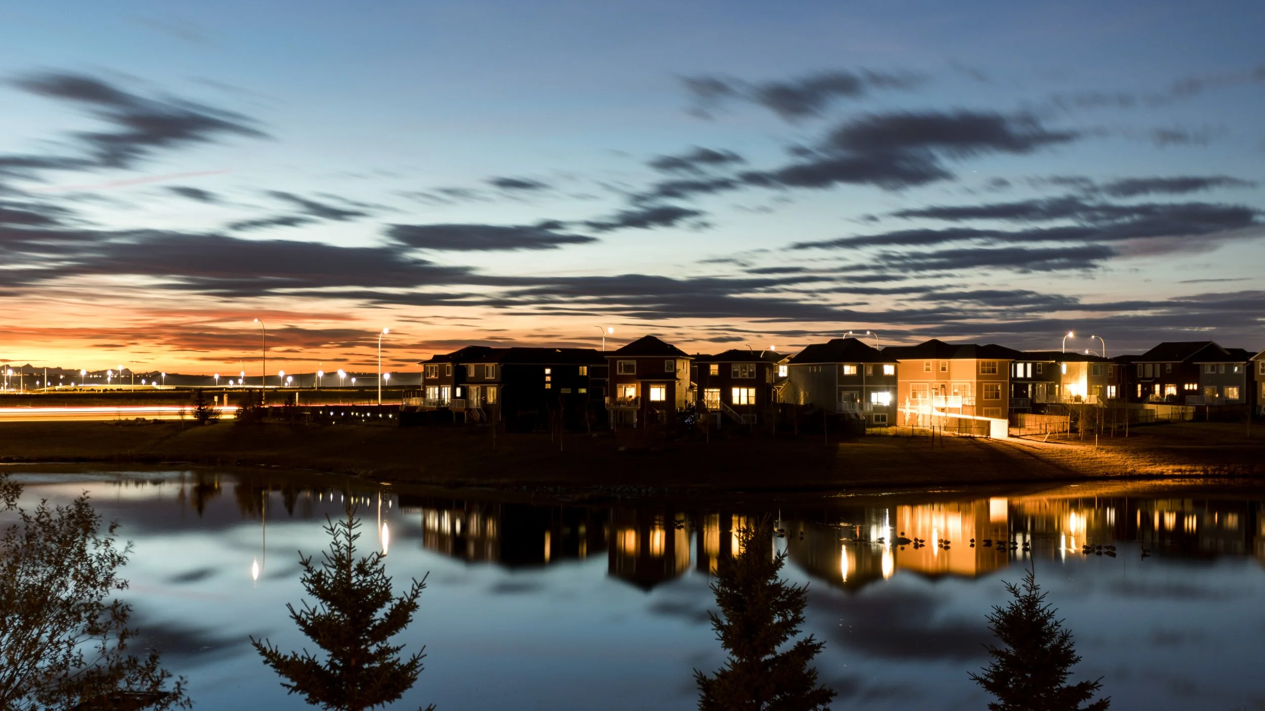 A lakeside neighborhood at dusk with illuminated houses and streetlights reflected in the calm water, and a colorful sky with clouds overhead.