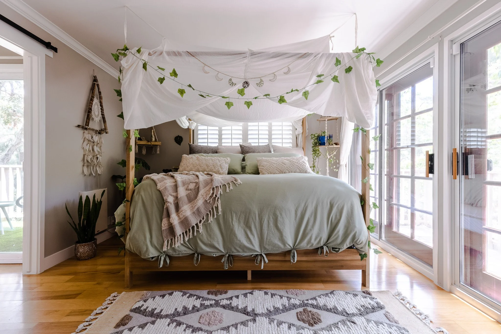 A cozy bedroom with a bed covered in light green bedding, decorated with pillows and a plaid throw blanket. A canopy of white fabric is draped above the bed with green ivy accents. Large windows and sliding glass doors let in natural light, with a pa