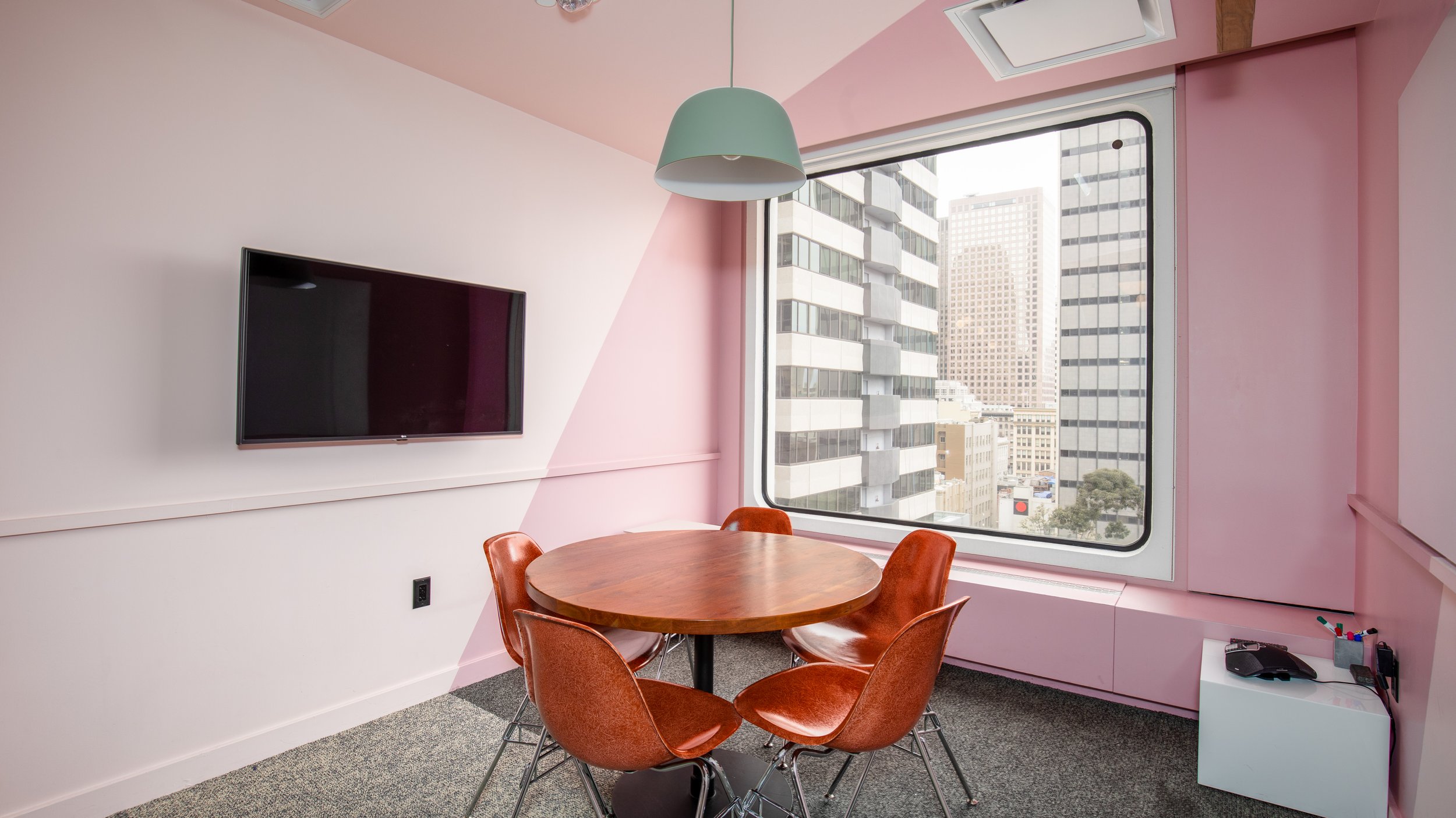 A meeting room with pink and white walls, a large window showing city buildings, a round wooden table with five orange chairs, a wall-mounted TV, a green pendant lamp, and a small white cabinet with office supplies.