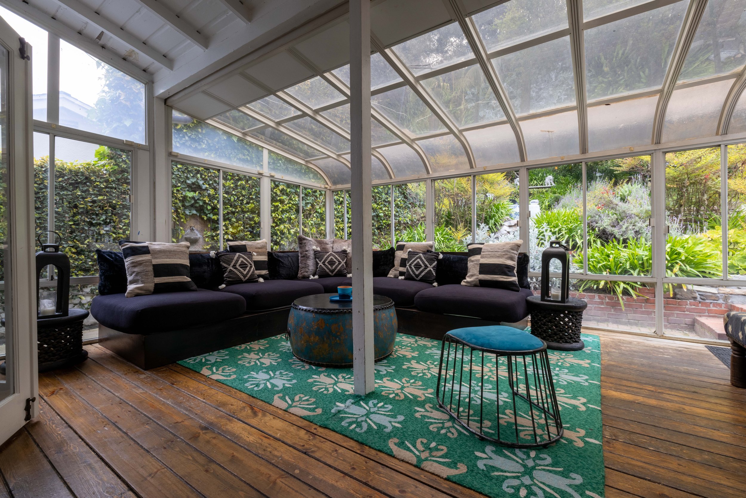 Interior of a sunroom or enclosed porch with black cushioned sectional sofa, assorted decorative pillows, a round blue coffee table, a teal and white patterned rug, and a small metal-framed stool. Glass windows reveal greenery and a garden outside.