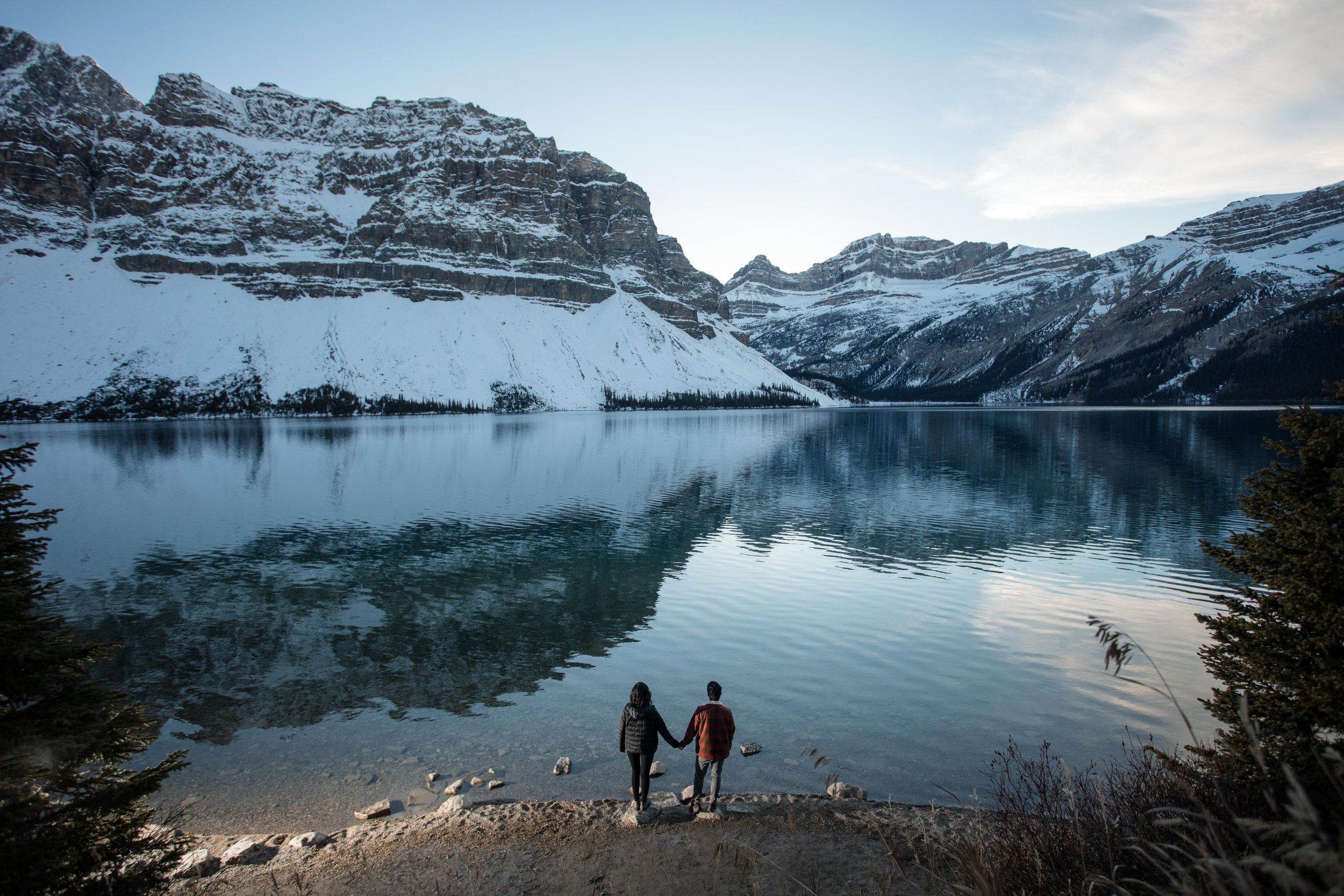 A couple holding hands by a lake with snow-covered mountains in the background.