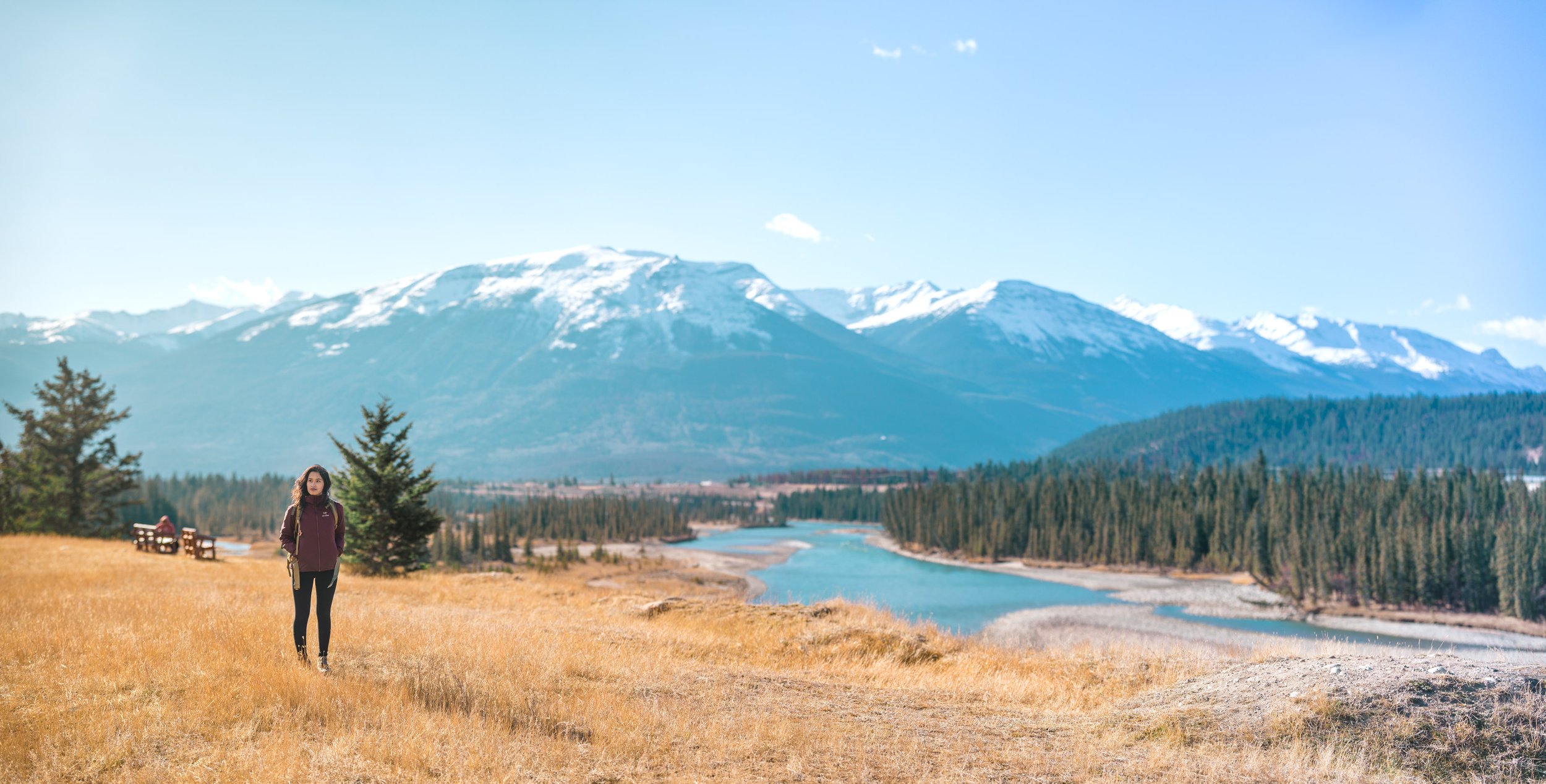 A woman walking through a grassy field near a river with snow-capped mountains in the background.