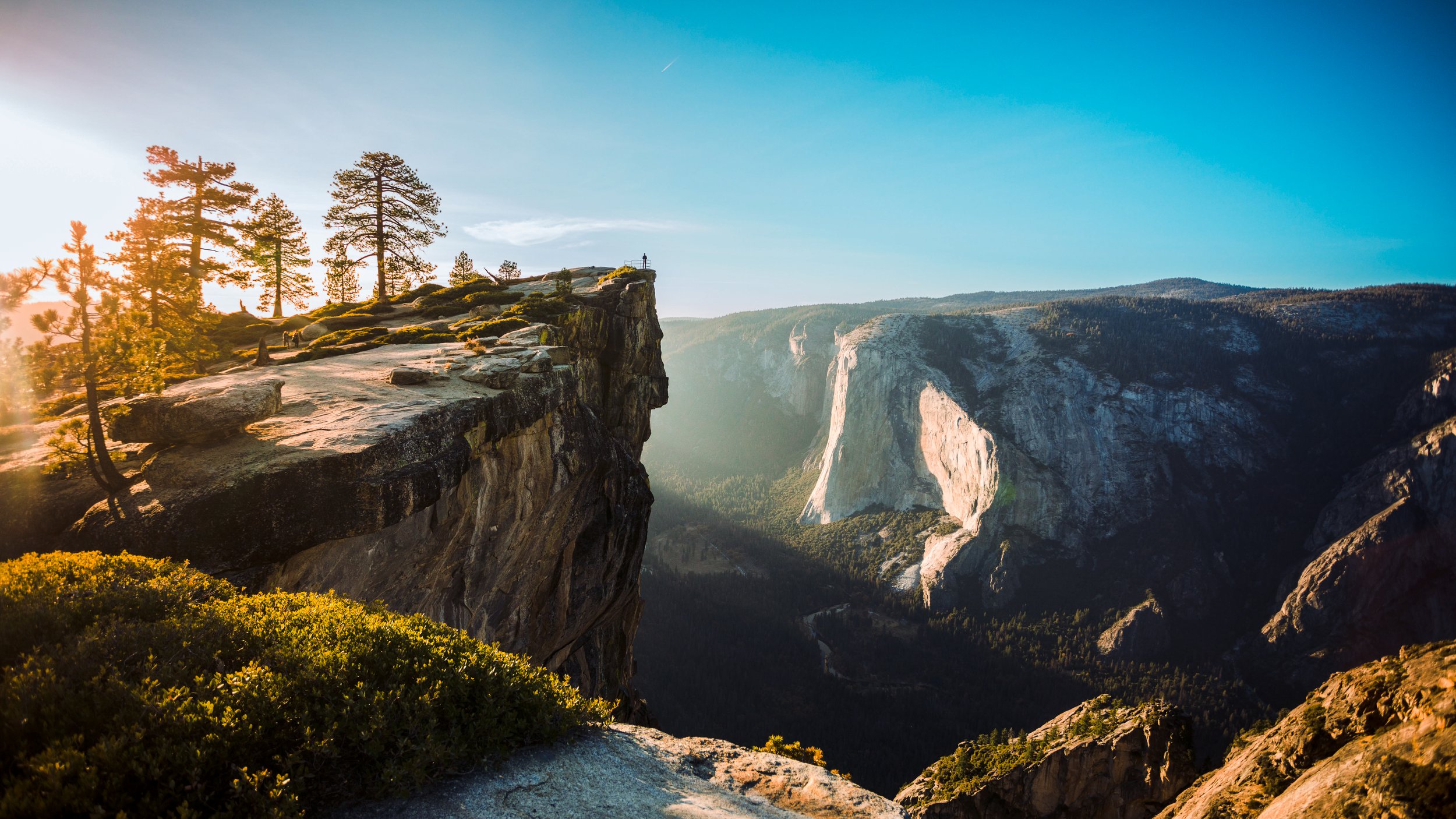 A scenic landscape of Yosemite National Park with a person standing on a cliff edge during sunset, overlooking the valley and towering cliffs.
