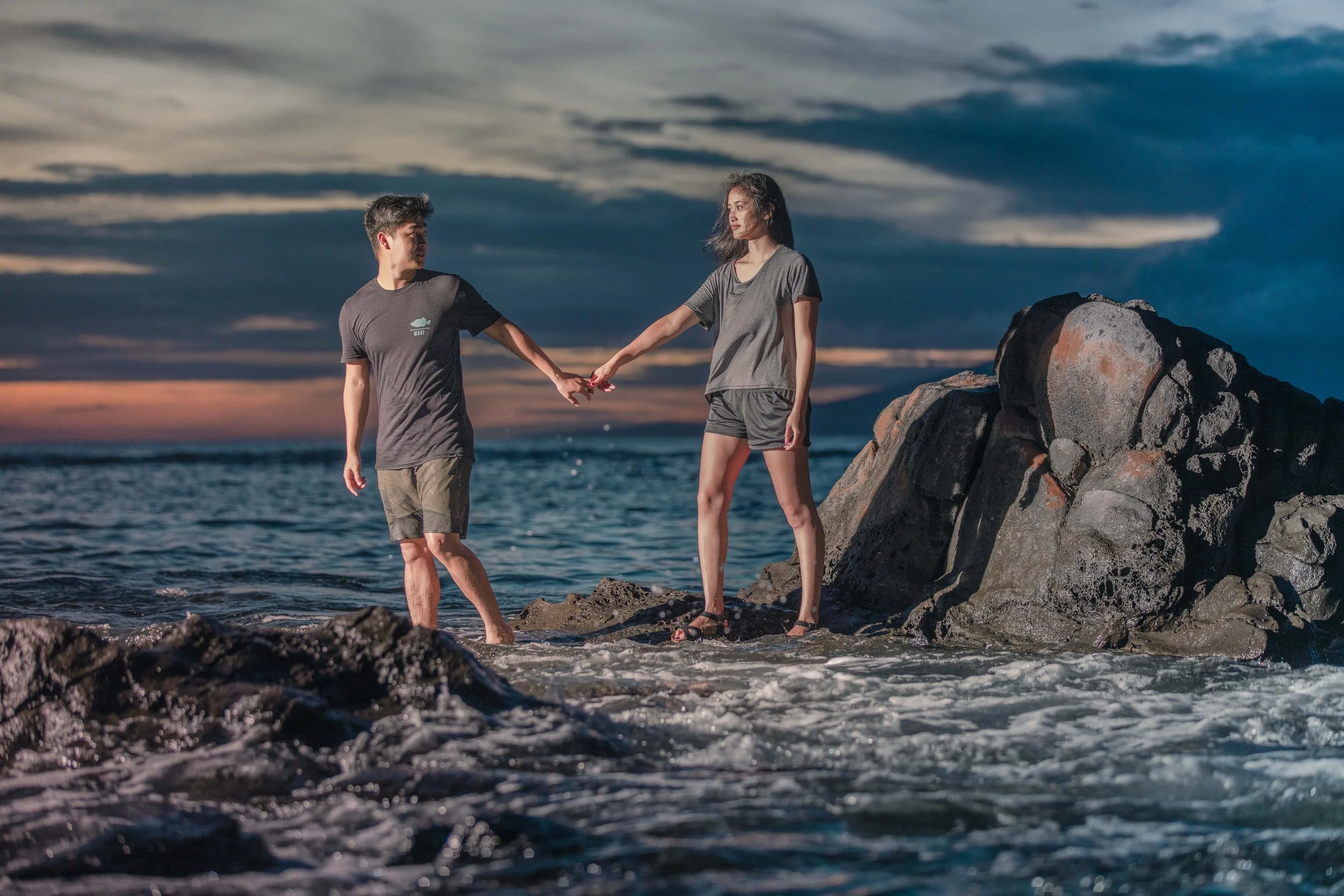 A young man and woman are holding hands on rocks at the beach during sunset, with dark clouds and the ocean in the background.
