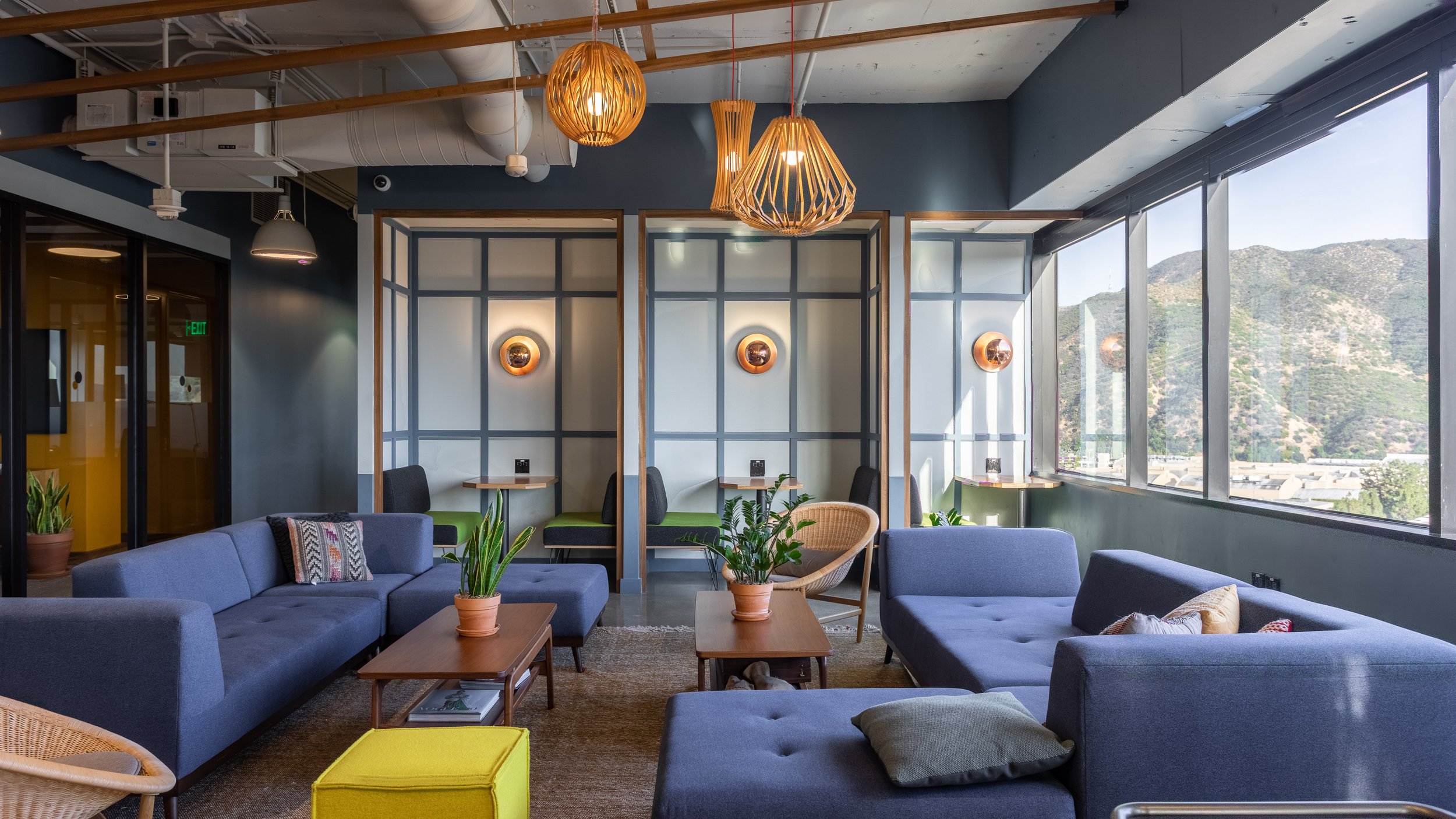 Modern lounge area with blue sofas, potted plants, wooden coffee tables, and private booths along the wall, featuring large windows with mountain views and woven pendant lights.