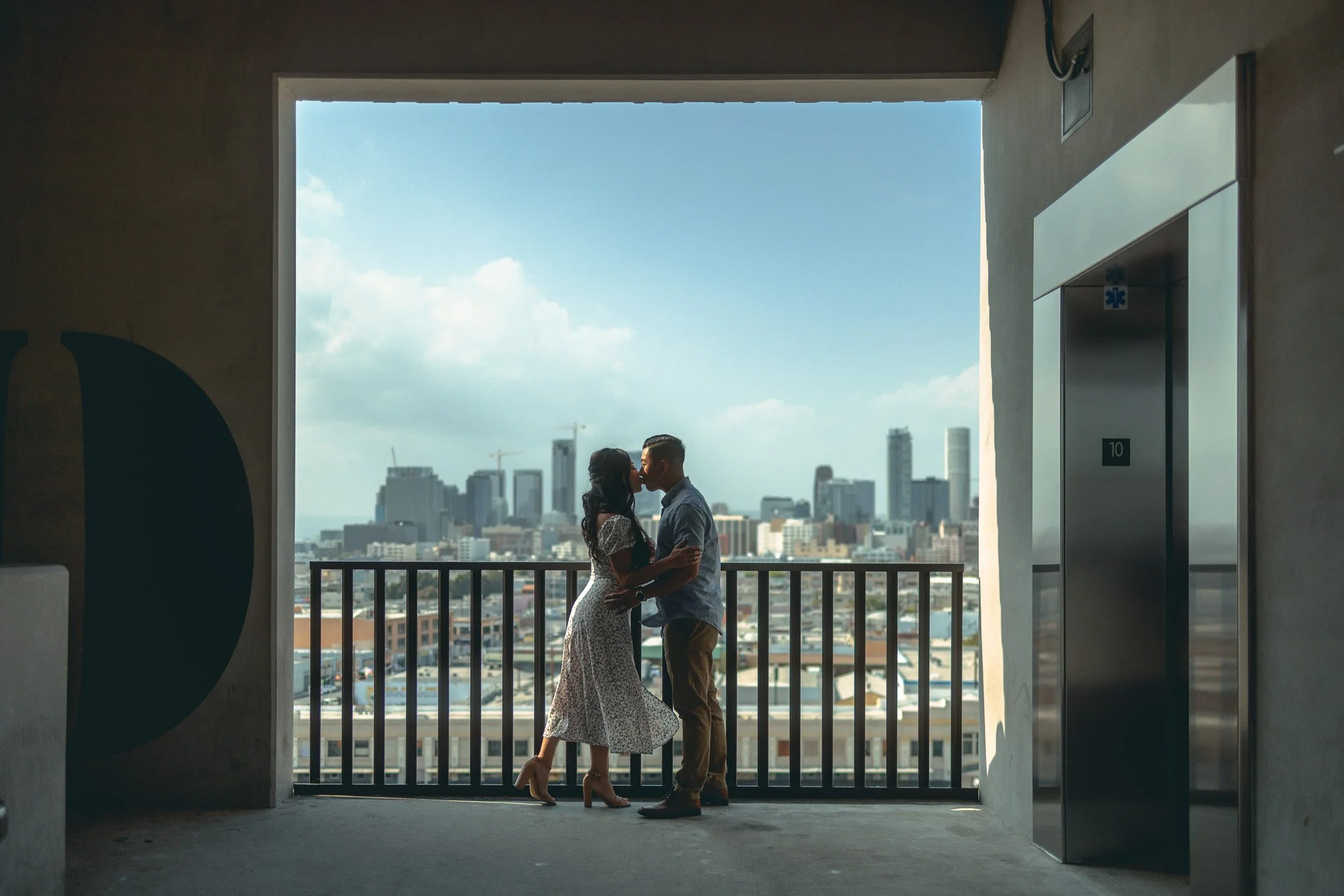 A couple sharing a kiss on a city balcony, with a skyline of tall buildings and skyscrapers in the background.