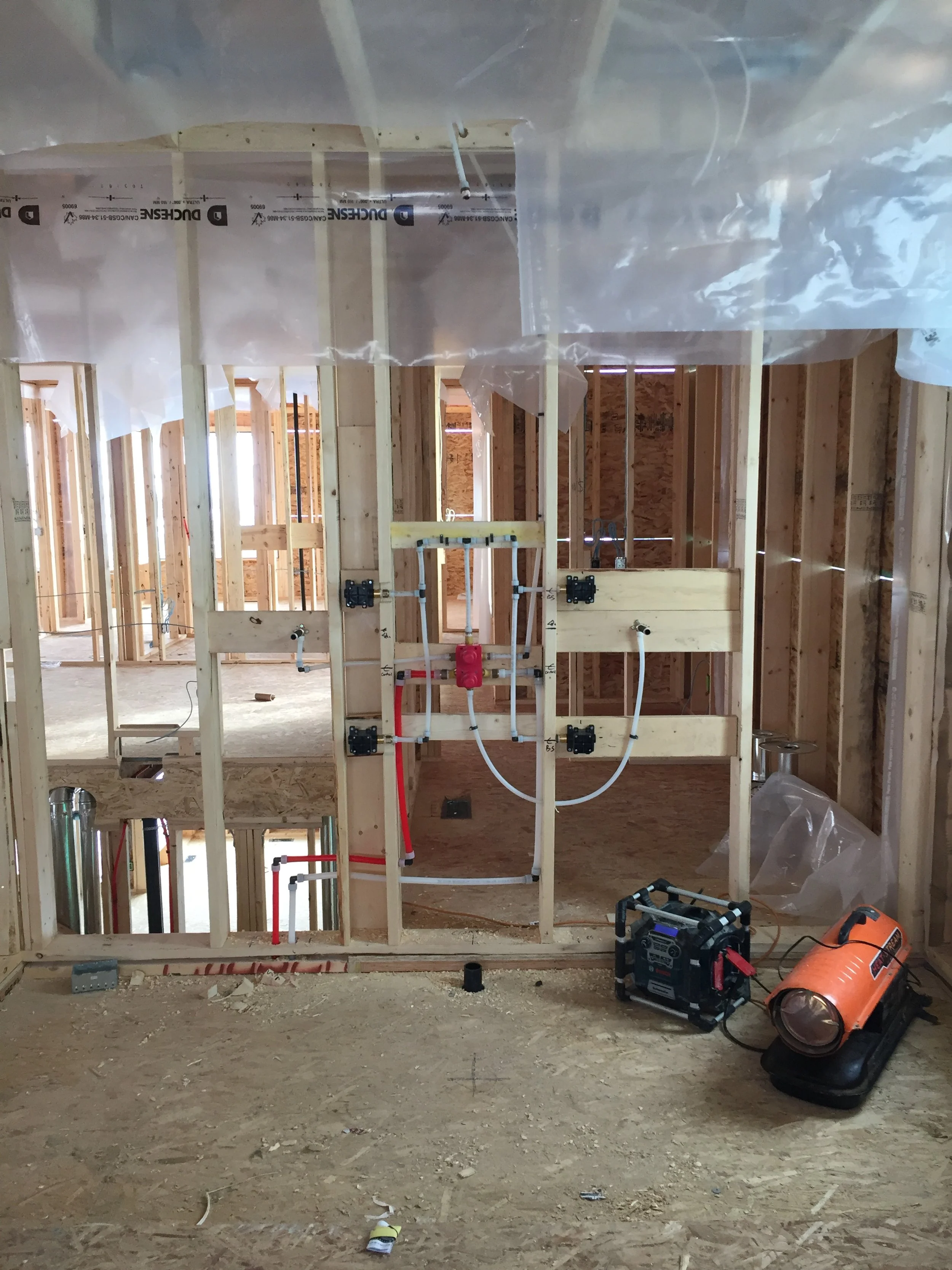 Interior view of a house under construction with exposed wooden framing, plumbing pipes, and insulation materials. Construction equipment is on the floor.