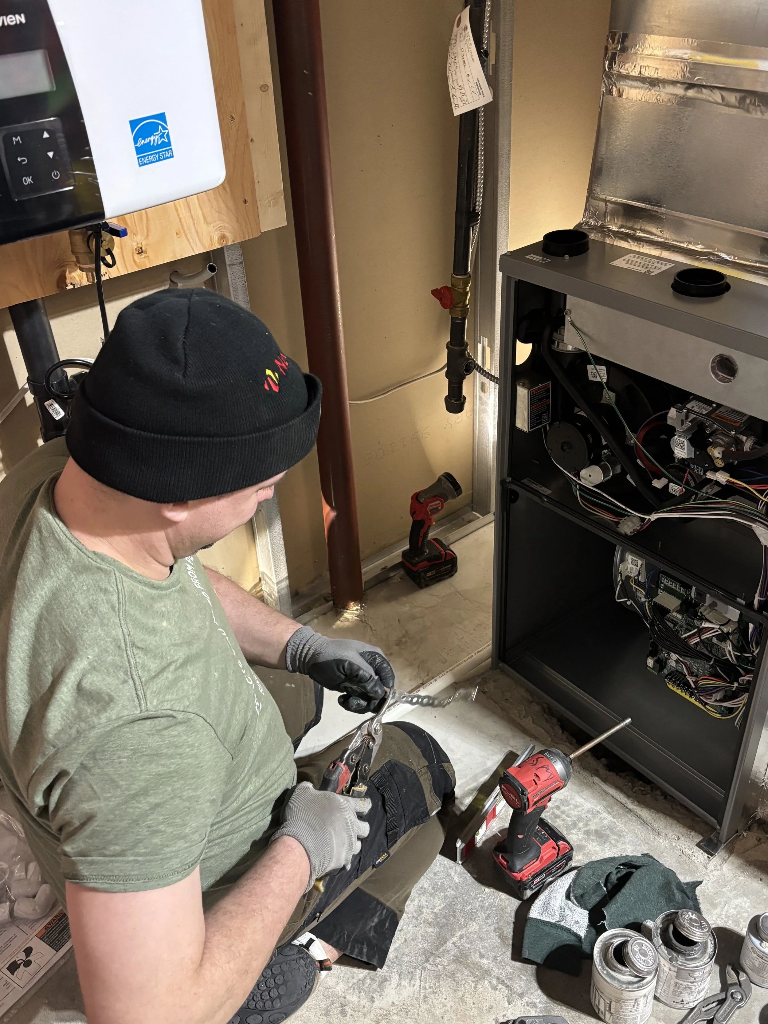 Steve Moore working on an electrical or HVAC unit, using a wrench, with tools and cans nearby in a maintenance room.
