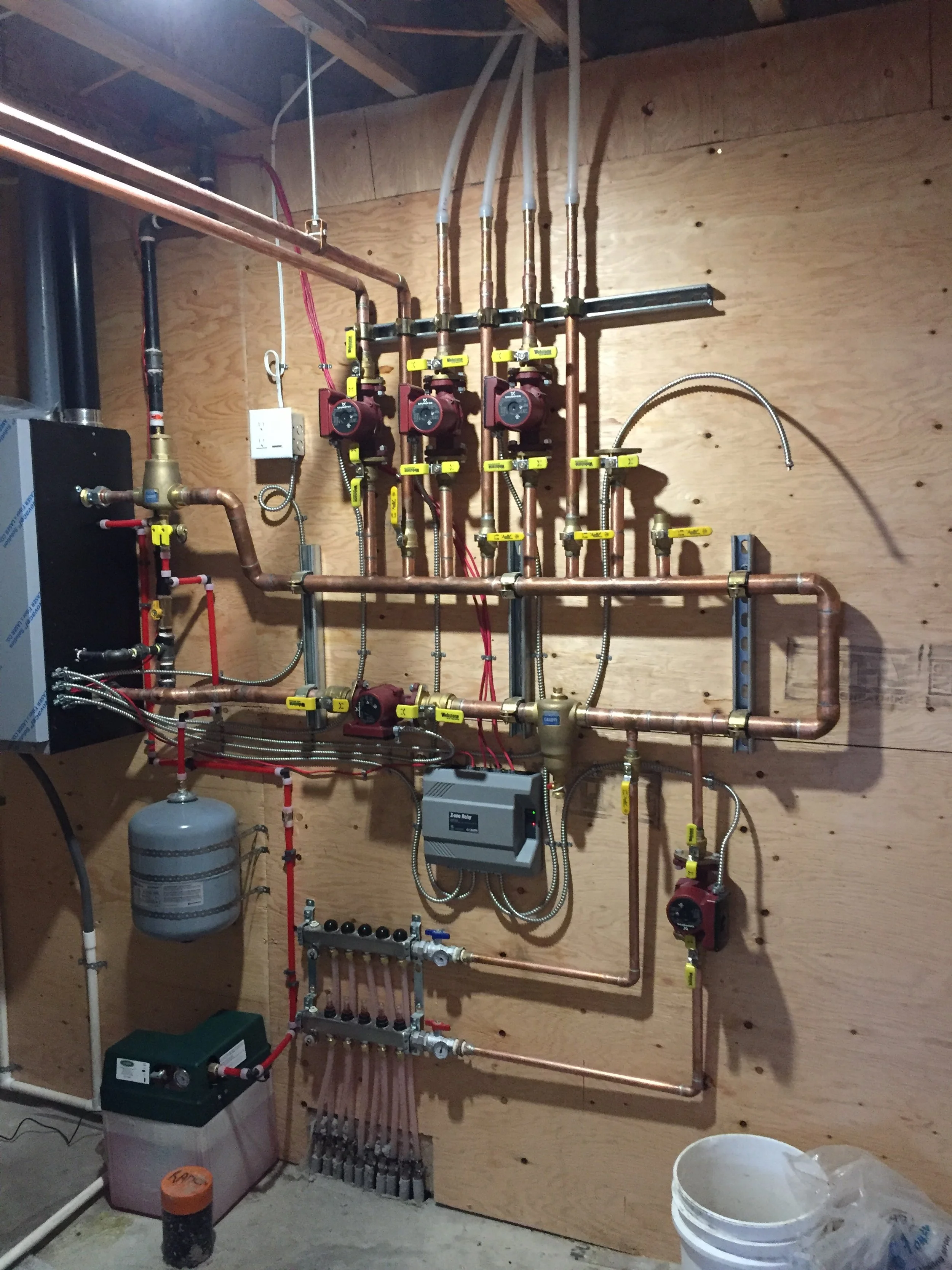 A plumbing and heating manifold system installed on a wooden wall, with copper pipes, pumps, valves, and a glycol tank.