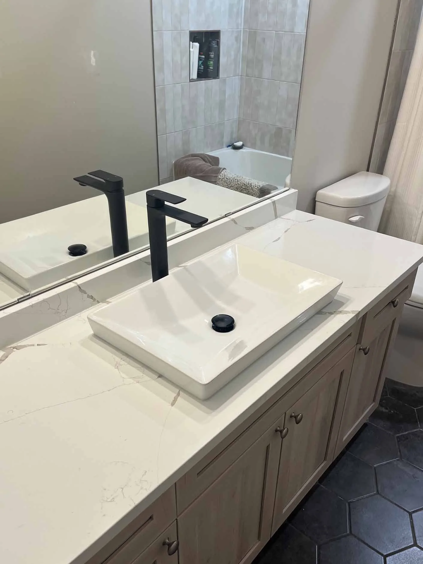 Bathroom with a white marble countertop, dual black faucets, rectangular vessel sinks, a large mirror, a toilet, a bathtub with a towel, and tiled walls.