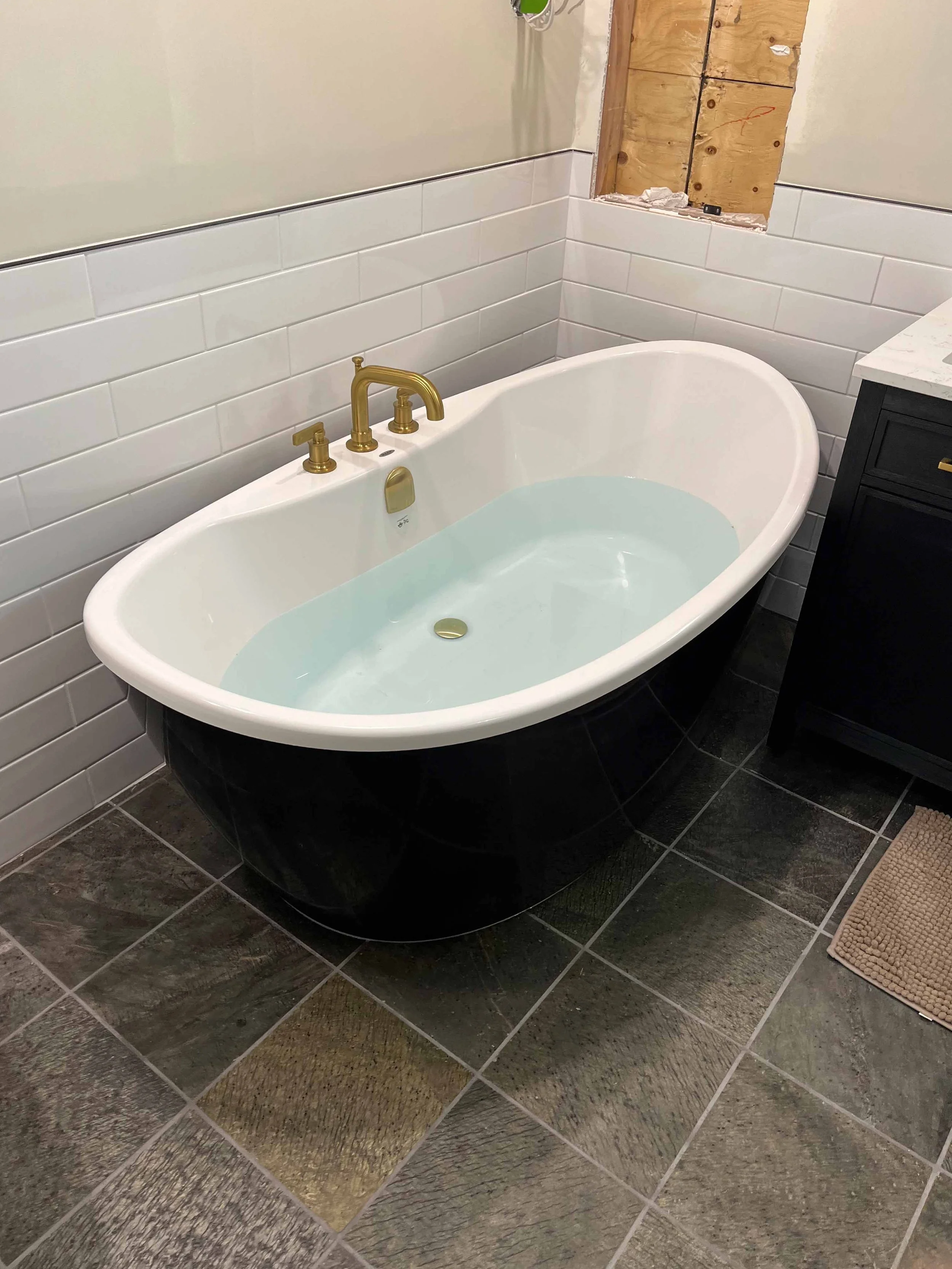 A modern bathroom with a freestanding bathtub filled with water, featuring gold faucets and fixtures. The tub is positioned next to a wall with white subway tiles and an unfinished section of wall. The floor is tiled with dark, textured tiles, and there is a black vanity with a white marble countertop nearby.