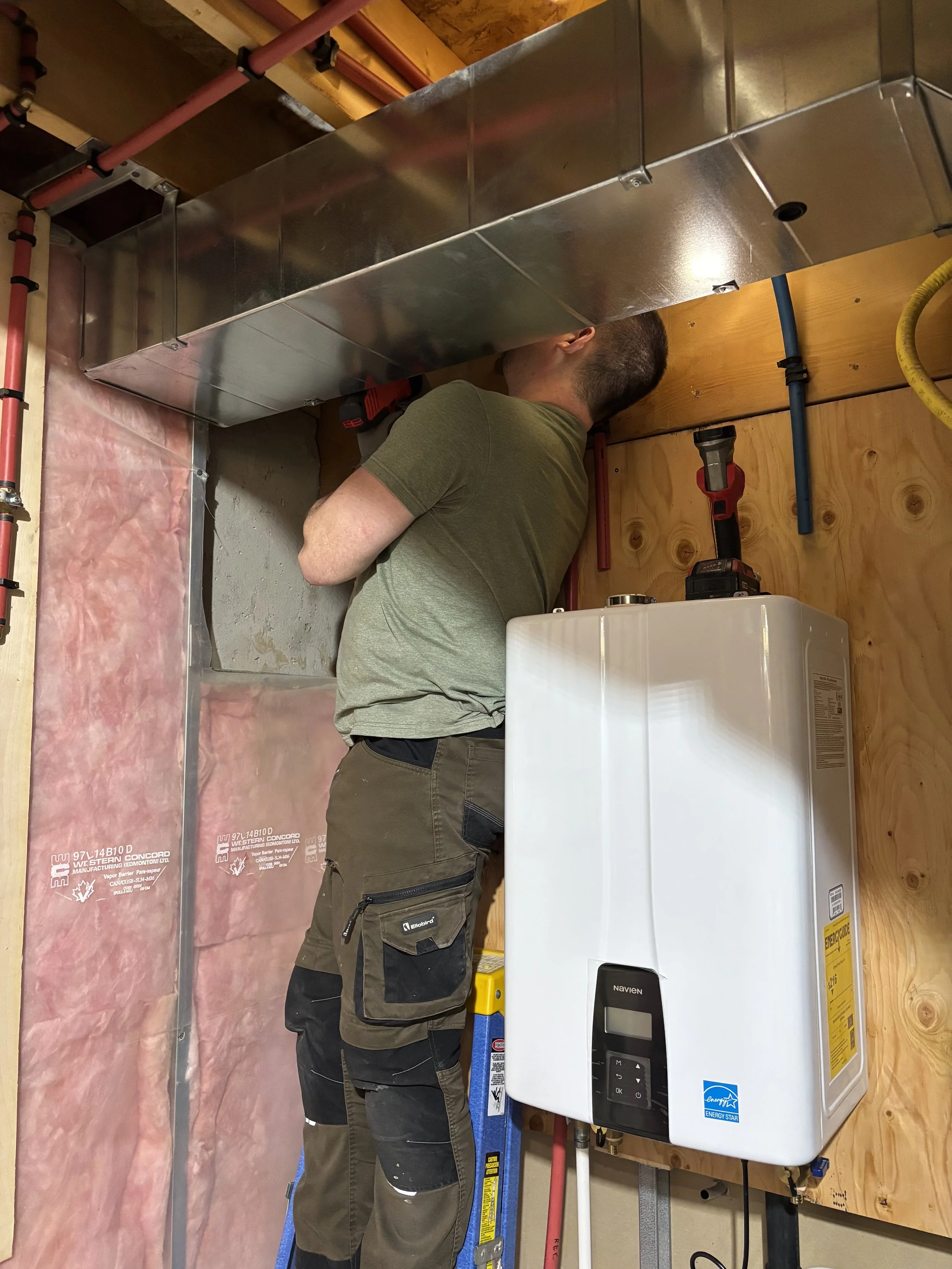 Steve Moore installing or repairing HVAC equipment in an unfinished basement or utility room with visible piping, insulation, and wooden paneling.