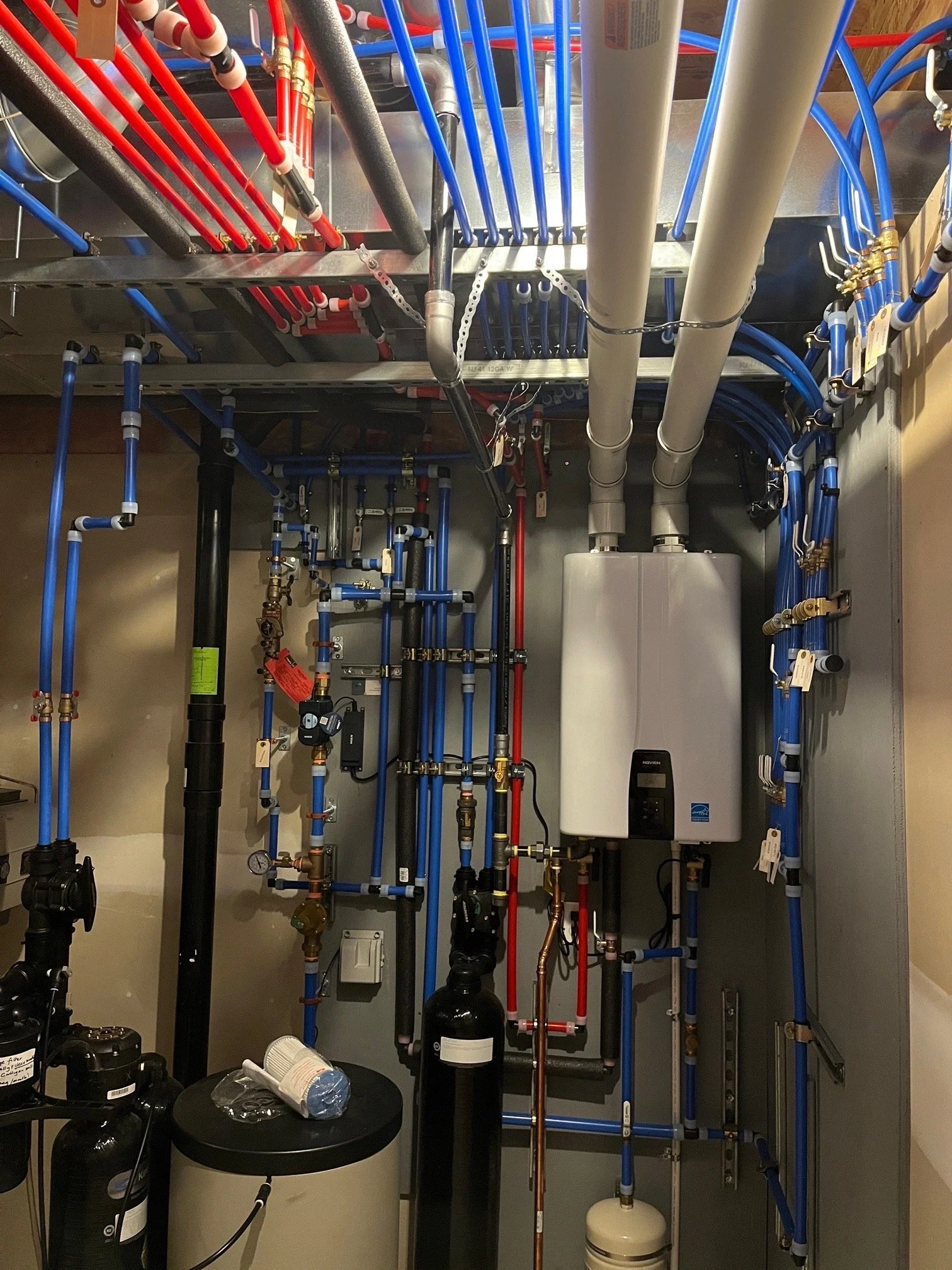 A utility closet with a network of blue, red, and gray pipes, a white tankless water heater, a black water filtration system, and various valves and gauges.