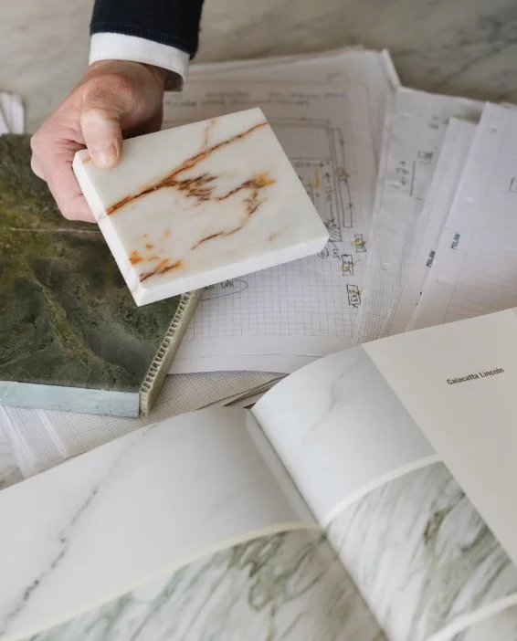 A hand holding a calacatta marble sample over architectural drawings and a green stone slab during a design consultation