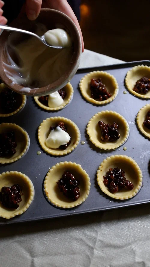 Spooning thick white cream cheese filling onto dark mincemeat inside raw pastry cases in a grey metal cupcake baking tray
